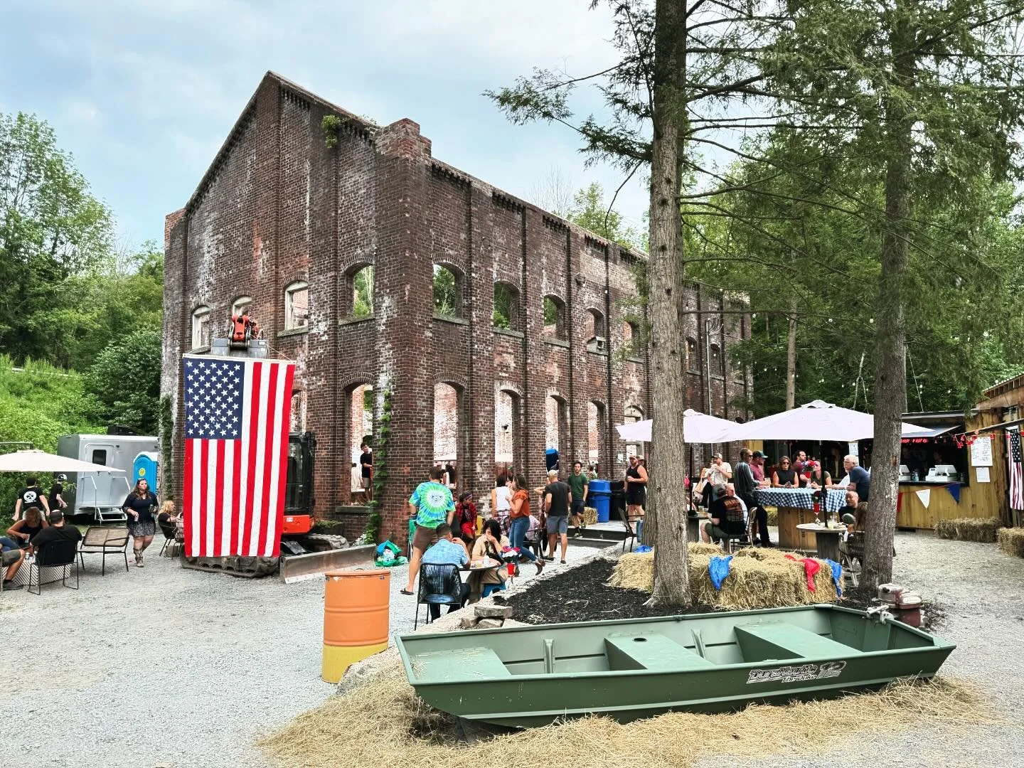 Outdoor gathering near a historic brick building with an American flag, people socializing, tables with umbrellas, hay bales, and a small green boat.