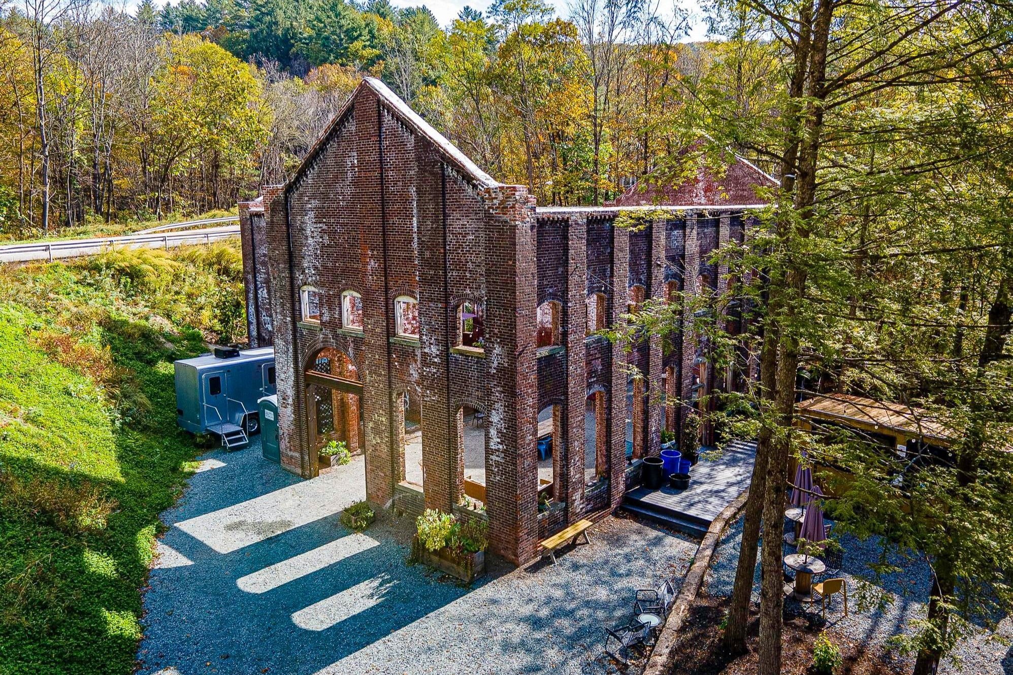 Abandoned brick building with open walls and missing roof in a wooded area.