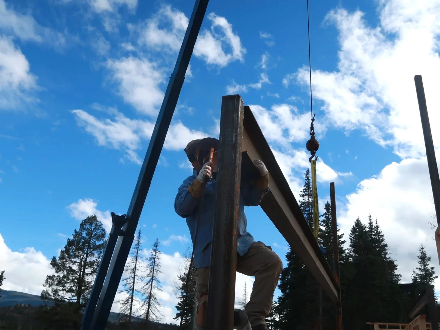 Construction worker wearing a helmet and gloves working on a steel beam outdoor under blue sky with clouds and trees in the background.