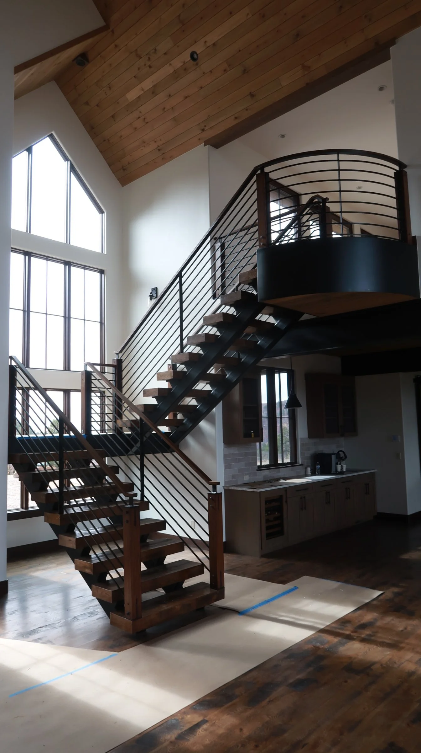 Interior view of a modern house with a wooden staircase leading to an upper level, large windows, a kitchen area with cabinets, a countertop, a window, and a wooden ceiling.