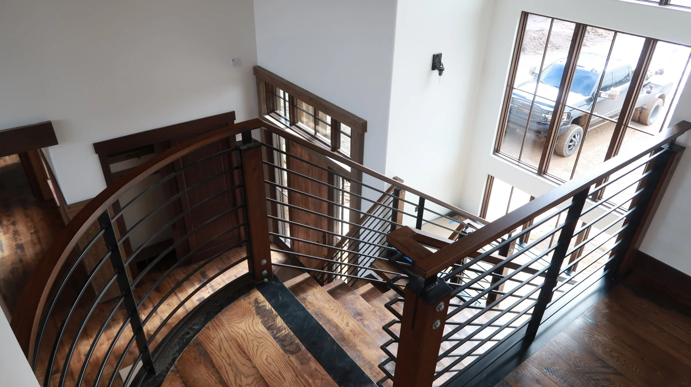 View from a staircase looking down, showing wooden flooring, metal railing, and large windows with a truck parked outside.