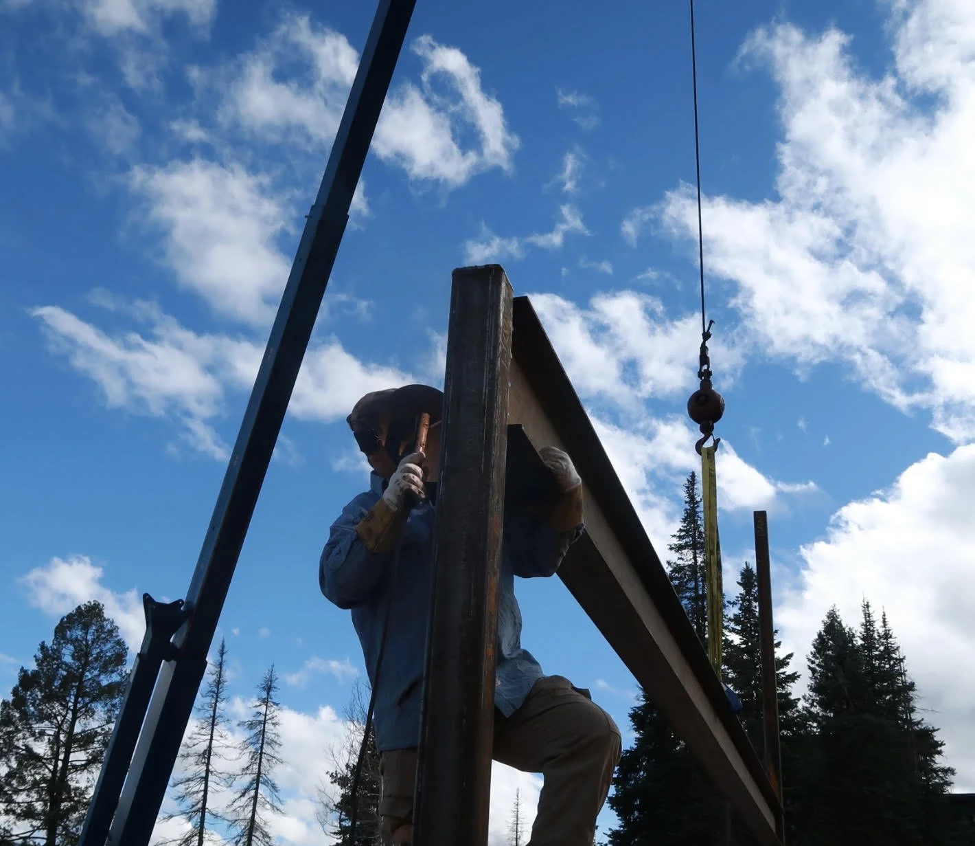 A person wearing gloves and a hat working on a wooden structure outdoors with a background of trees and a partly cloudy sky.
