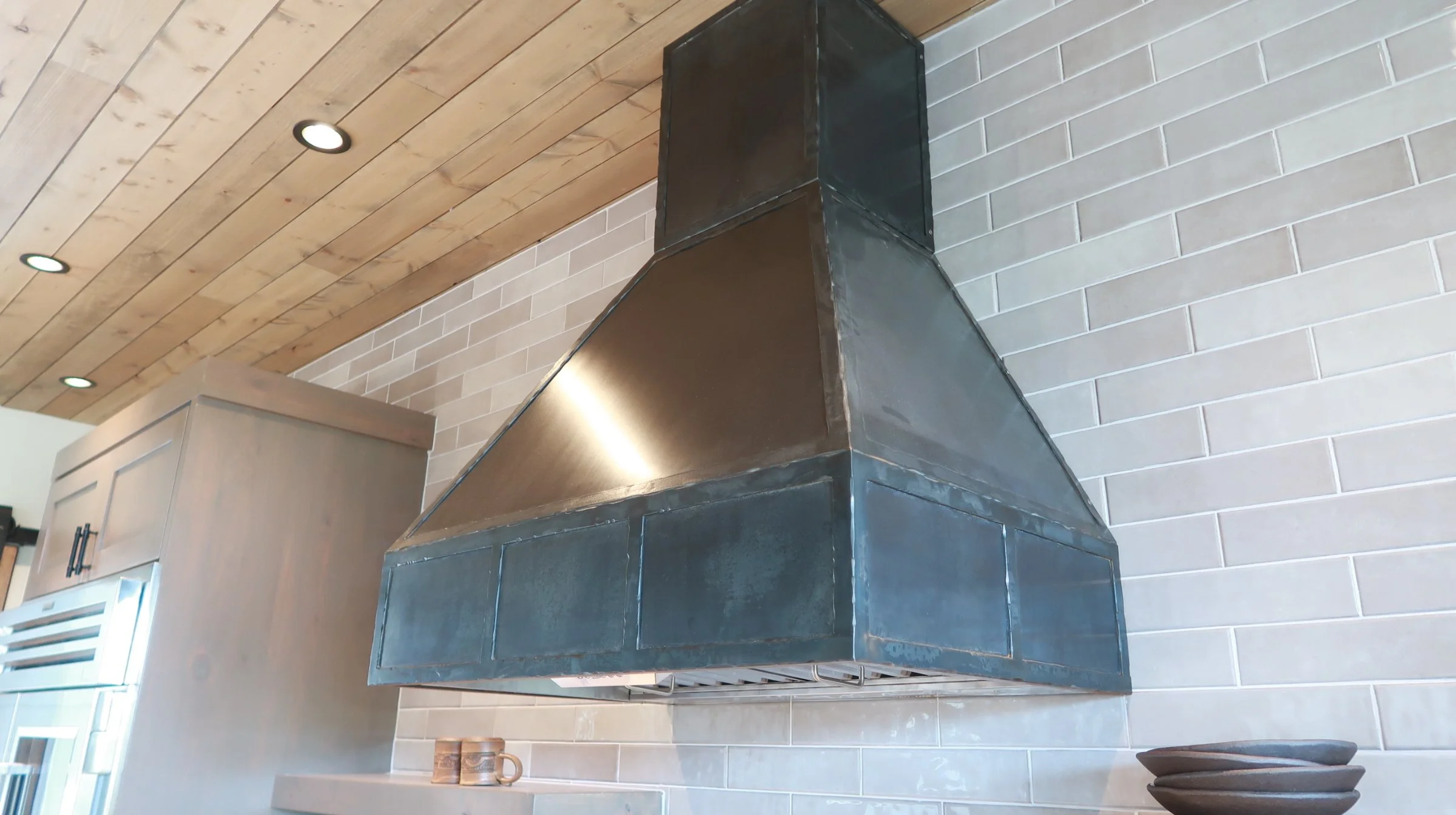 A stainless steel range hood mounted above a kitchen stove, against a beige brick backsplash with a wooden ceiling featuring recessed lighting.