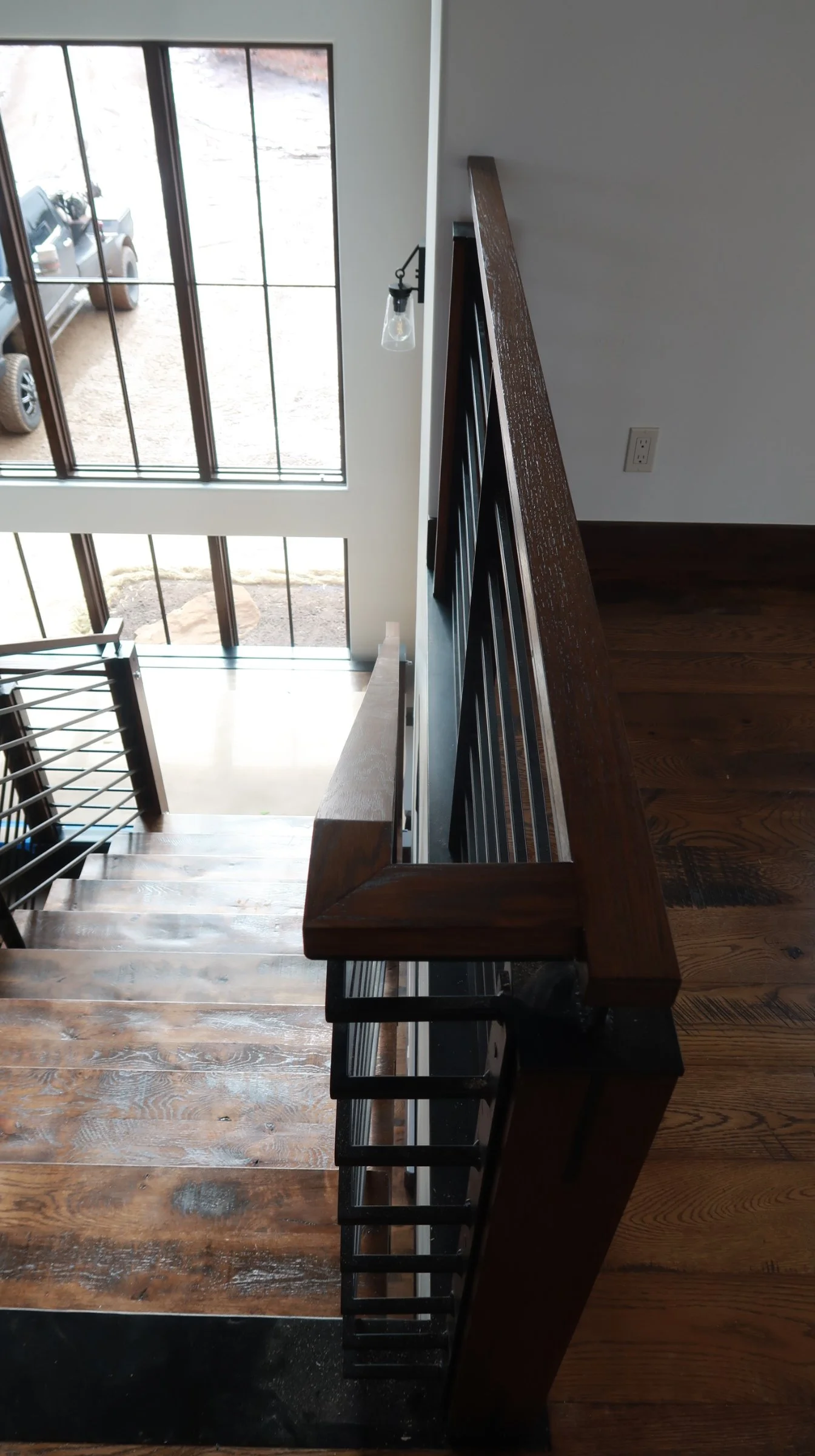 View of a staircase with wooden steps and black metal railing, from the top floor looking down towards large windows and a parking lot outside.