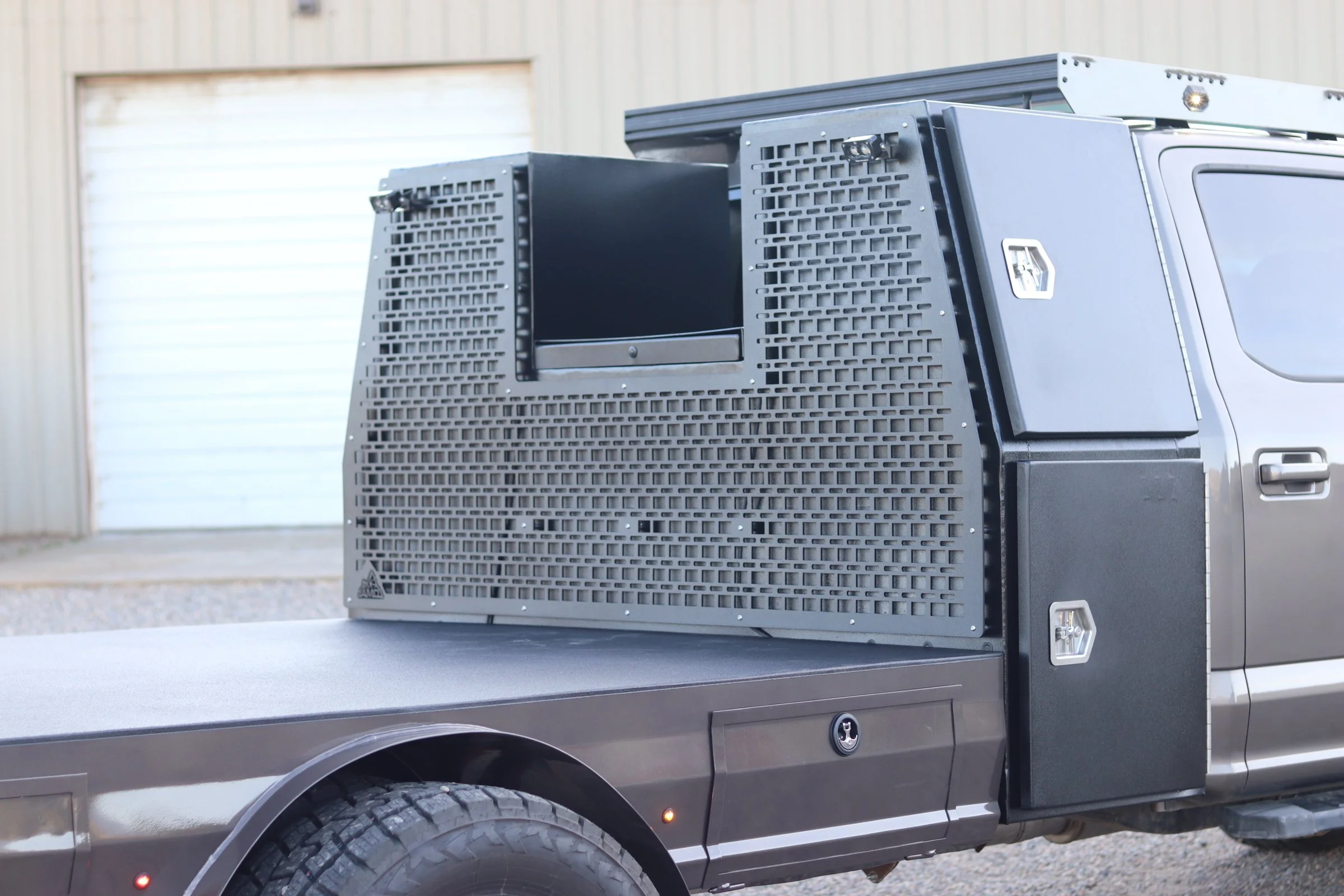 A black armored vehicle with a large, rectangular, ventilated shield on the back, parked outside a building with a metal roll-up door.