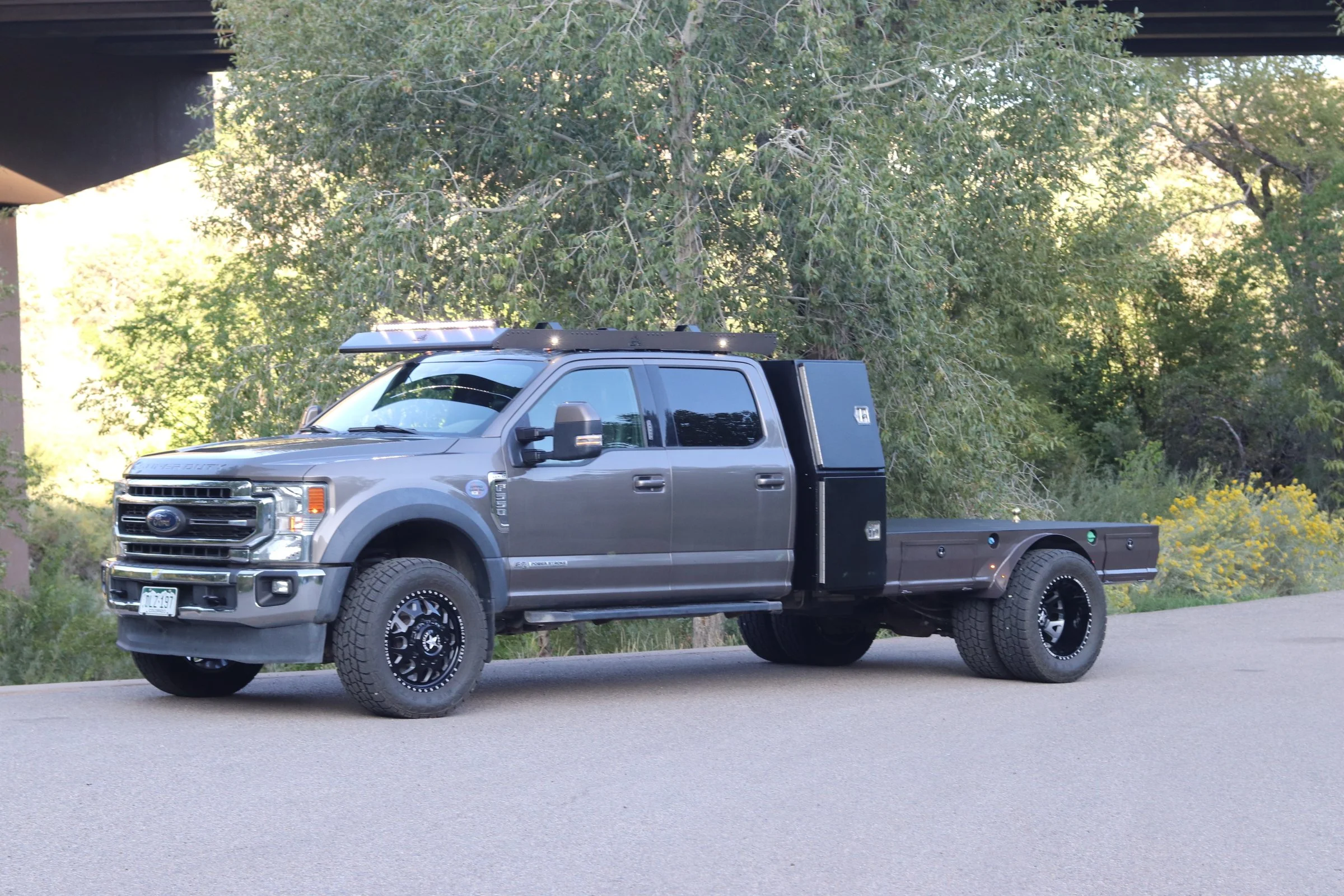 A gray Ford F-150 truck with custom black wheels and off-road tires, parked on an asphalt road with green trees and yellow flowers in the background.