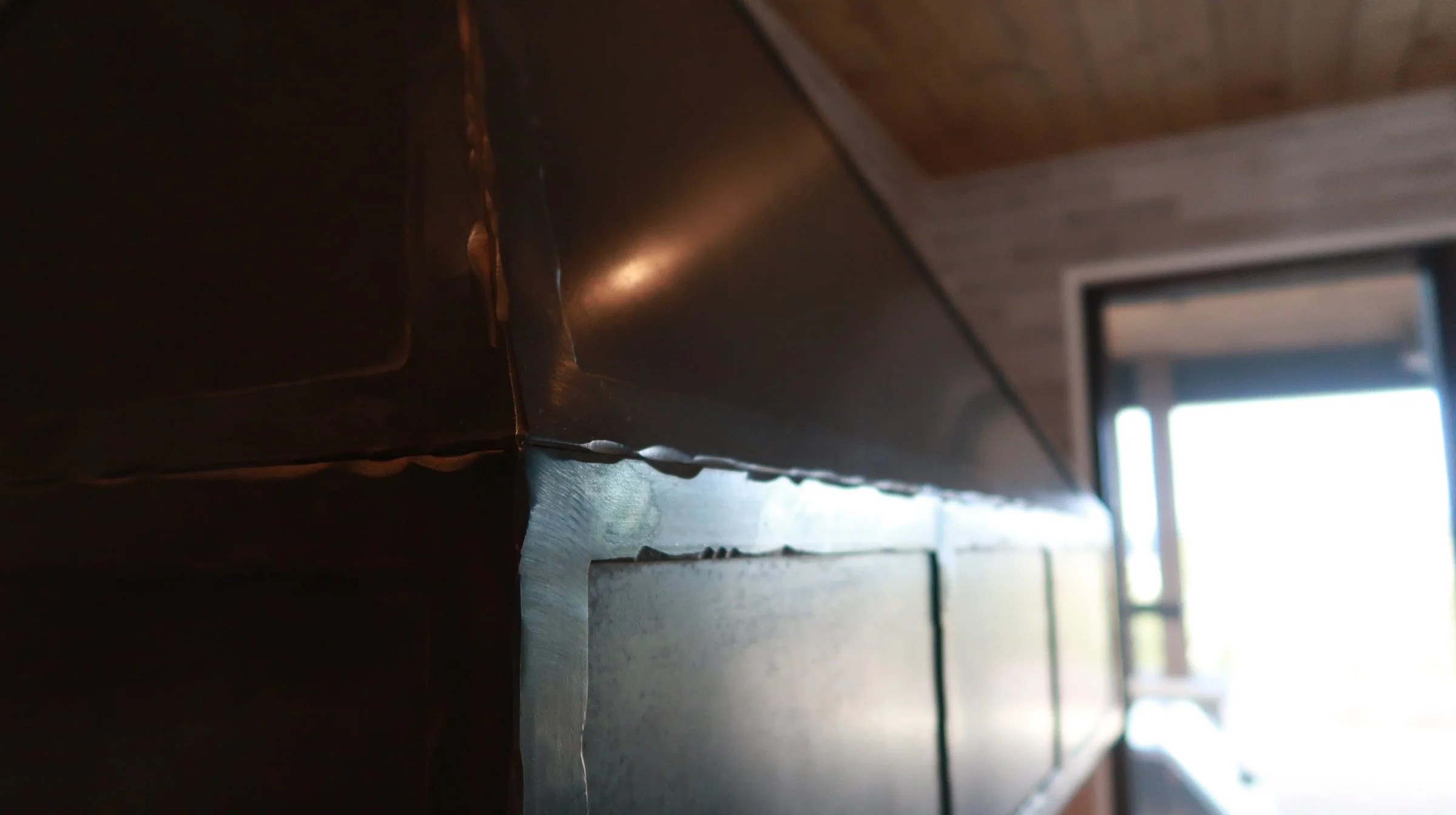 Close-up of a dark wooden cabinet or shelf with visible joints, in a room with a wooden ceiling and a large window or glass door in the background, allowing natural light to illuminate the space.