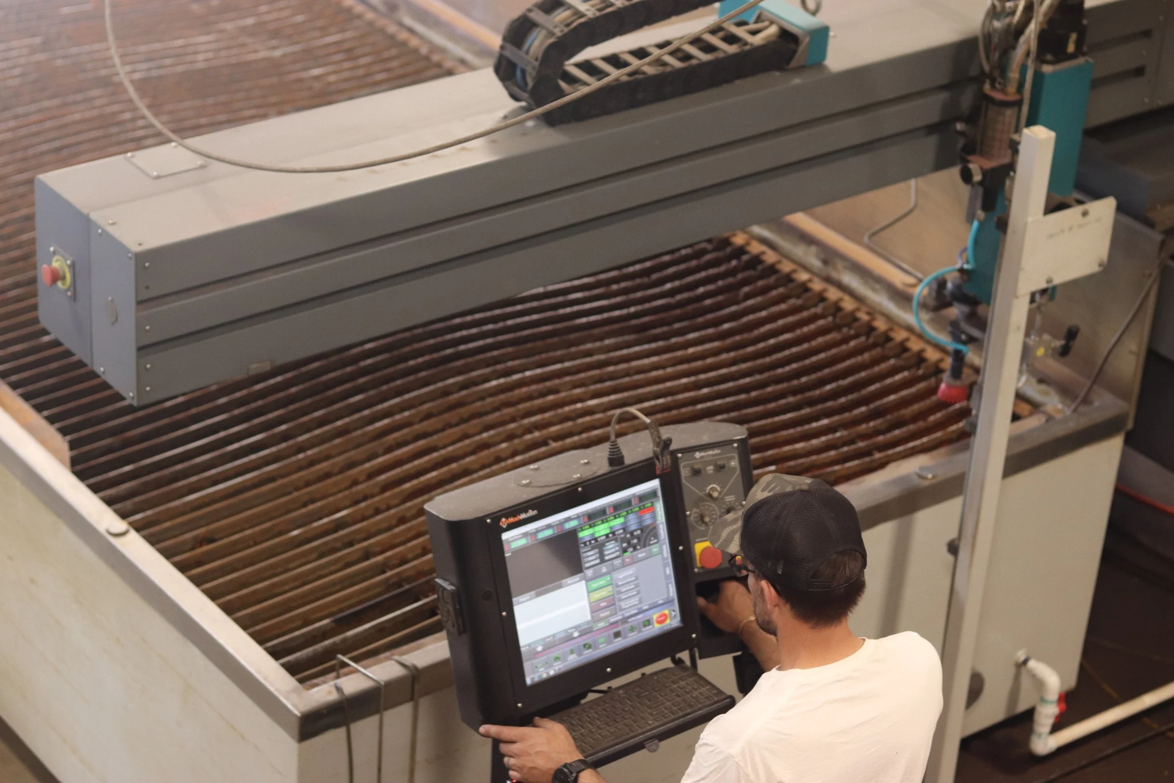 A person operating a computer with a large monitor and control panel, overseeing a machine that appears to be a hydroponic or agricultural system with brown, water-filled ridges.