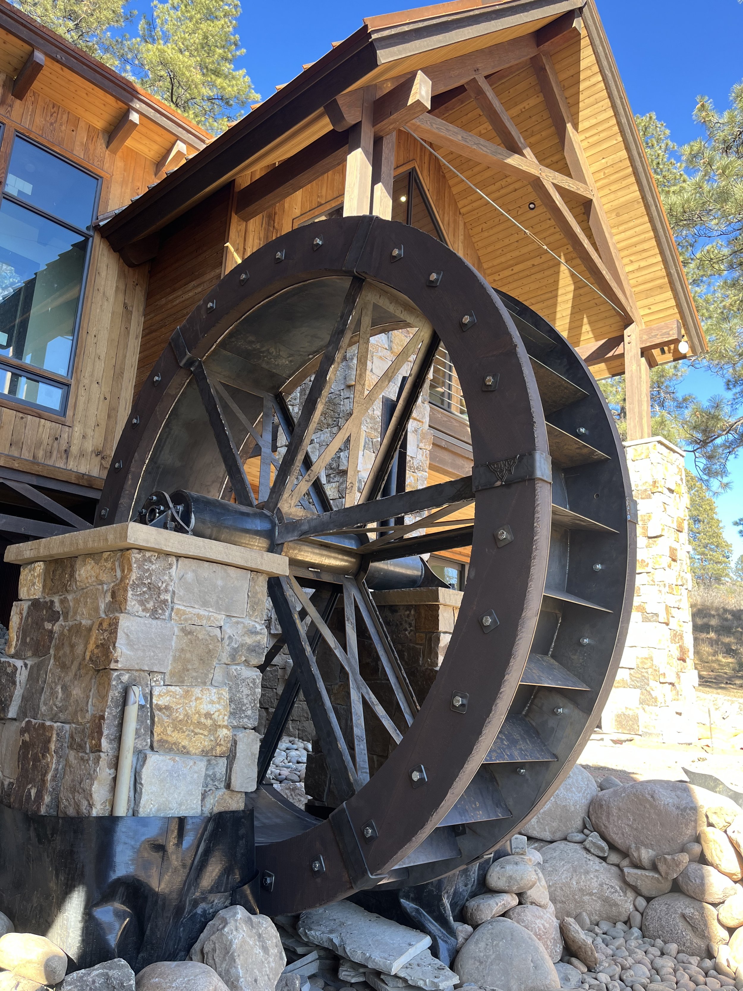 A large wooden waterwheel in front of a house with stone and wood exterior, surrounded by rocks and trees under a clear blue sky.