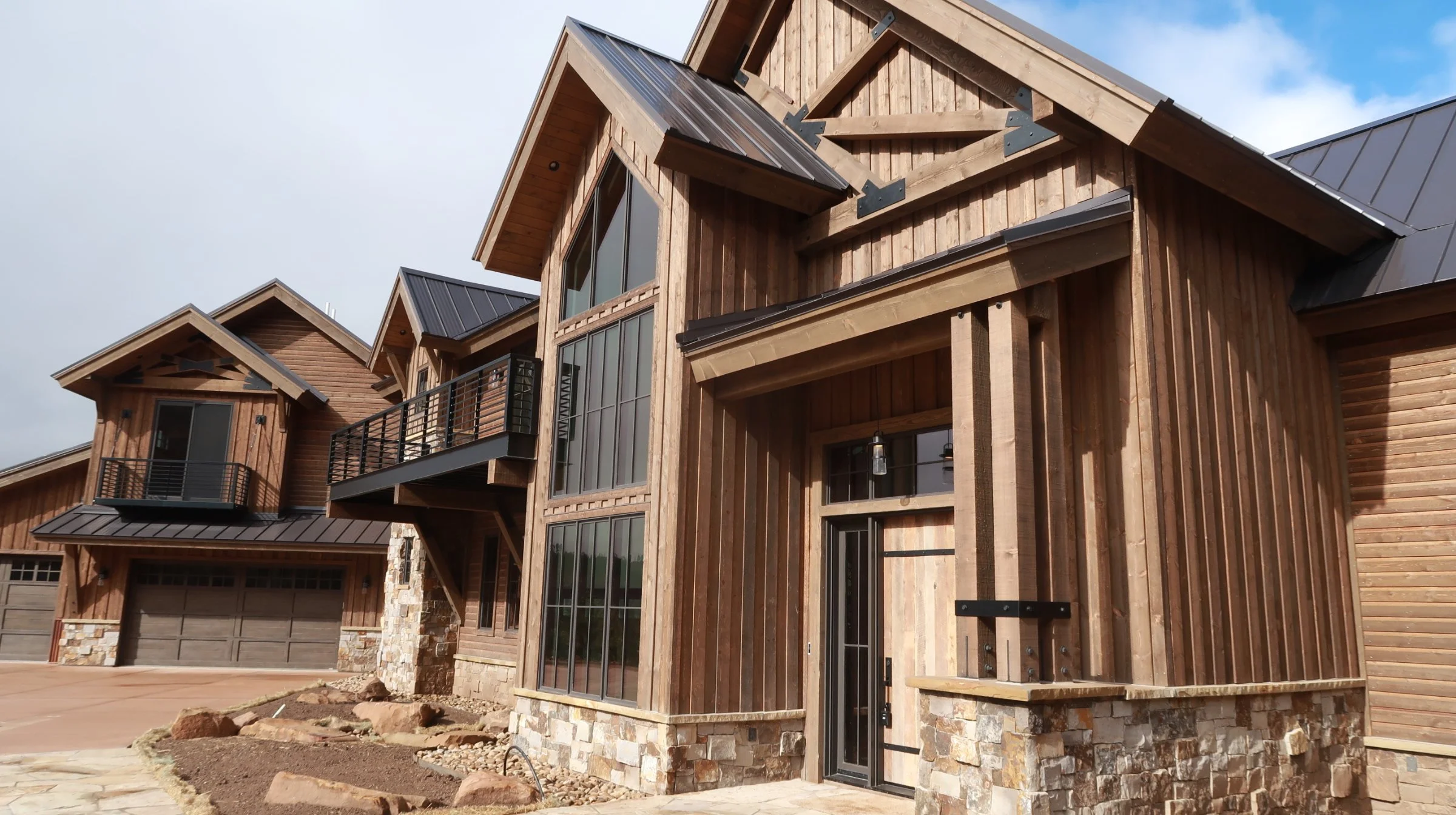 Modern multi-story house with wooden exterior, large glass windows, and stone accents, under a partly cloudy sky.