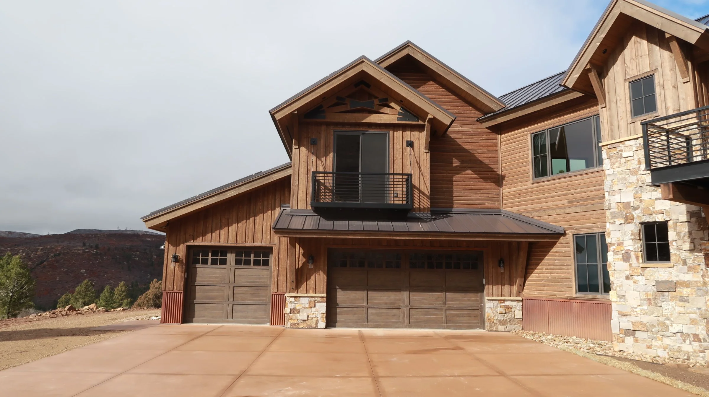 A modern house with wood siding, stone accents, and metal roofing, featuring a garage with dual doors, a small balcony with black railing, and large windows, set against a hillside landscape.