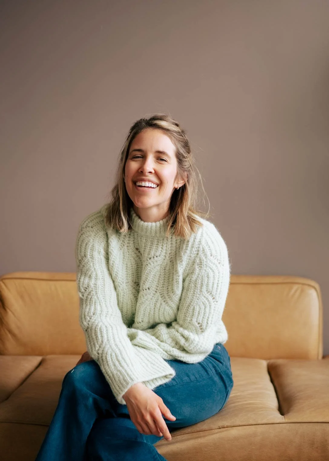 Abbey Kramer DDS with shoulder-length blonde hair smiling and sitting on a tan leather couch, wearing a cream cable-knit sweater and blue jeans.