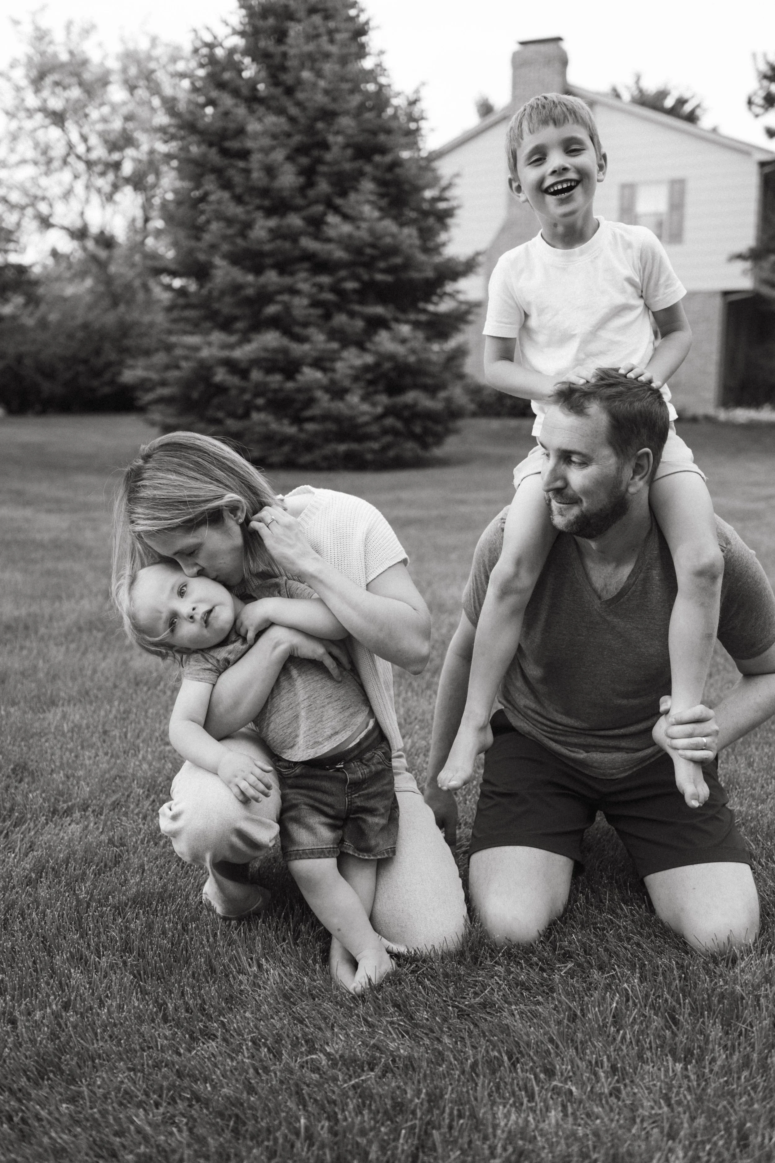 A black and white photo family of four in a grassy yard. The father is kneeling on the grass with a young boy sitting on his shoulders, both smiling. The mother is kneeling on the grass, kissing a young girl on the cheek, who is wrapped around her neck.