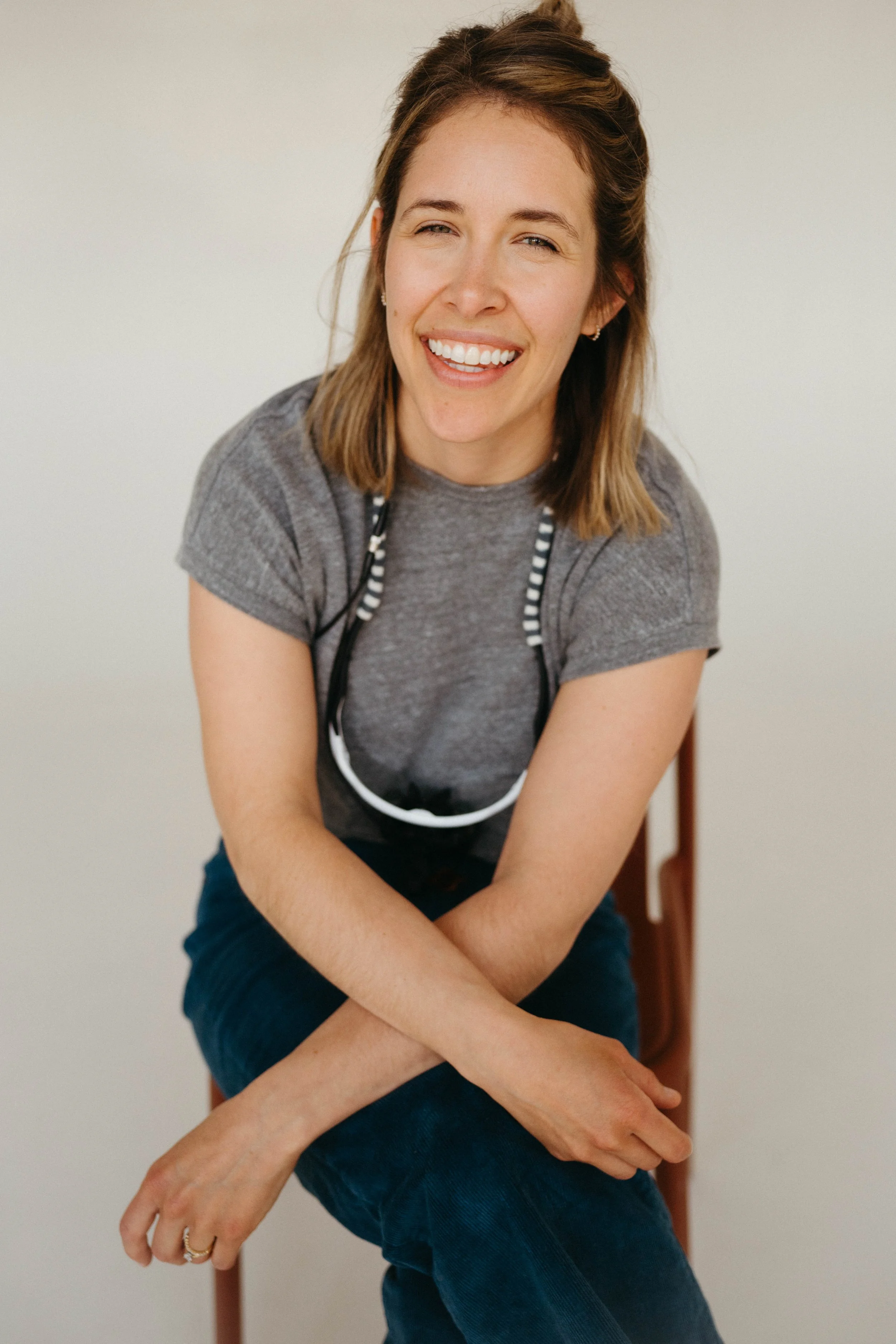 Abbey Kramer DDS with shoulder-length blonde hair, wearing a gray t-shirt and dark jeans, sitting on a chair, smiling and looking at the camera.