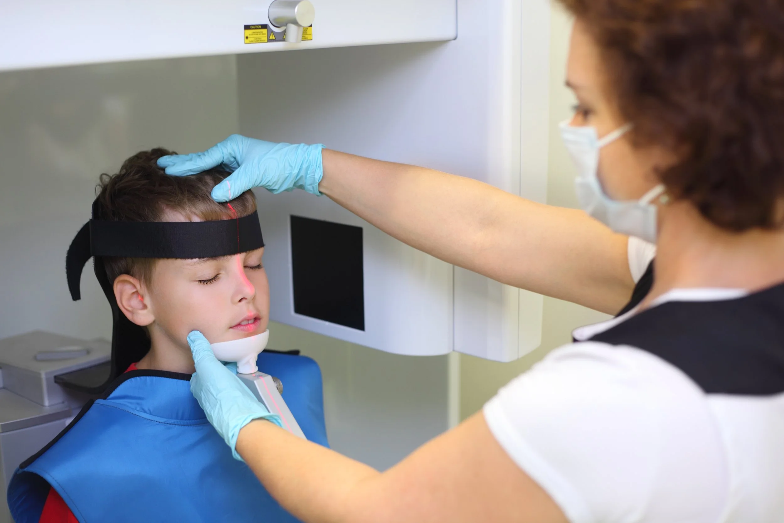 A healthcare professional wearing a face mask and gloves placing an alien headband on a young boy's head during a medical scan with a head coil machine.