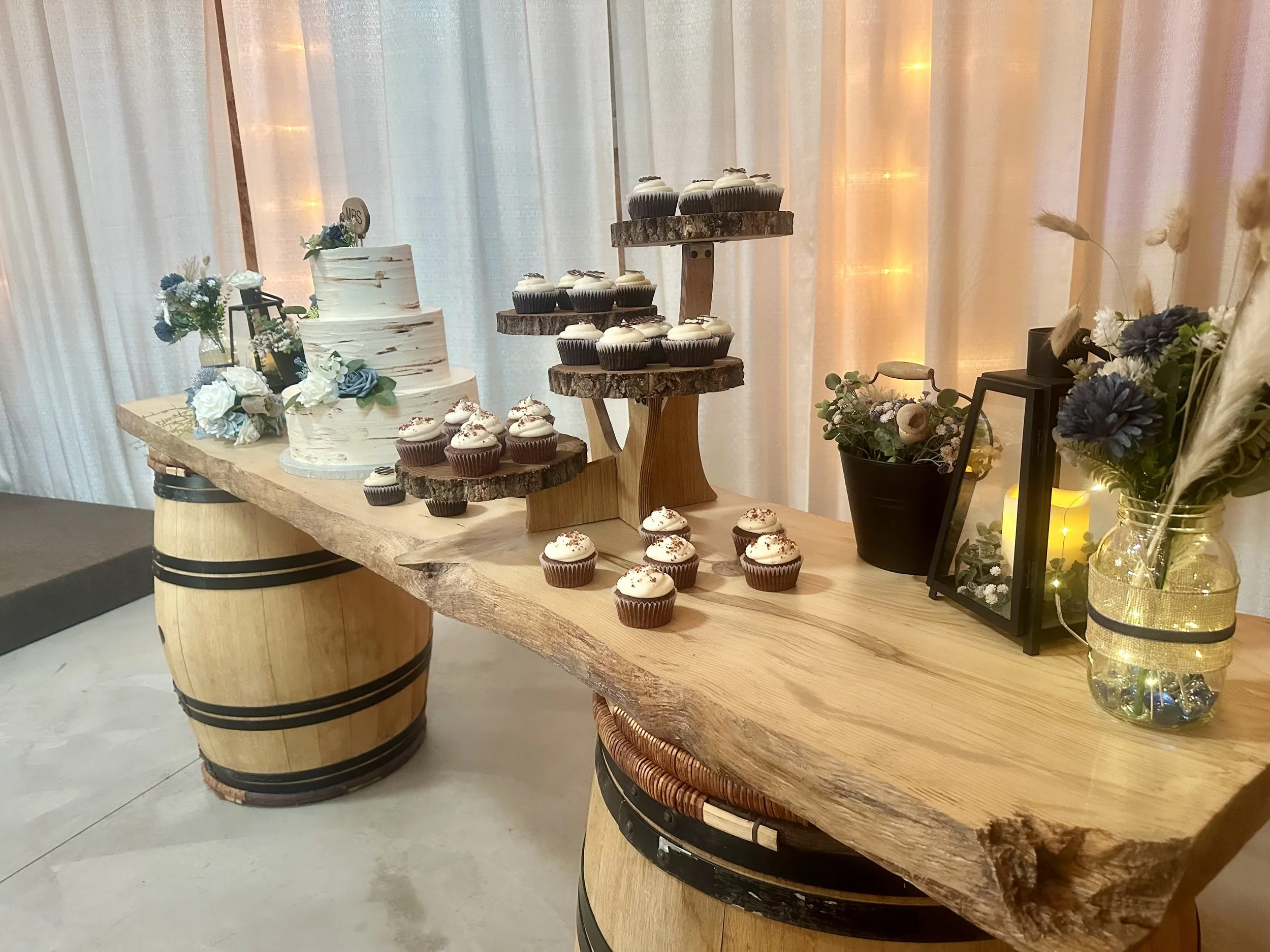 Wedding cake and cupcake display on live edge table with wine barrels