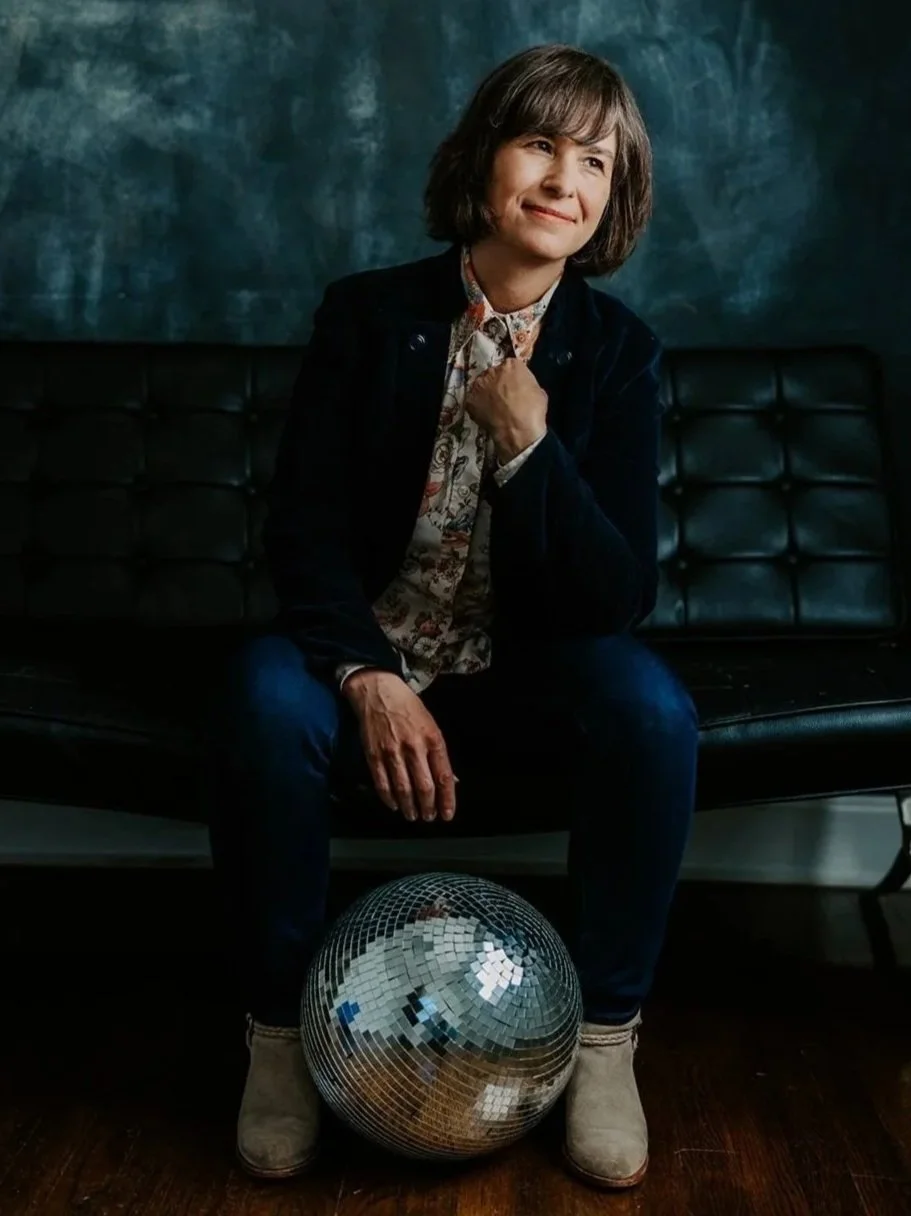 A woman with short brown hair sits on a black leather couch, wearing a floral blouse, dark jacket, and jeans, with a reflective disco ball between her legs and a blue textured wall background.
