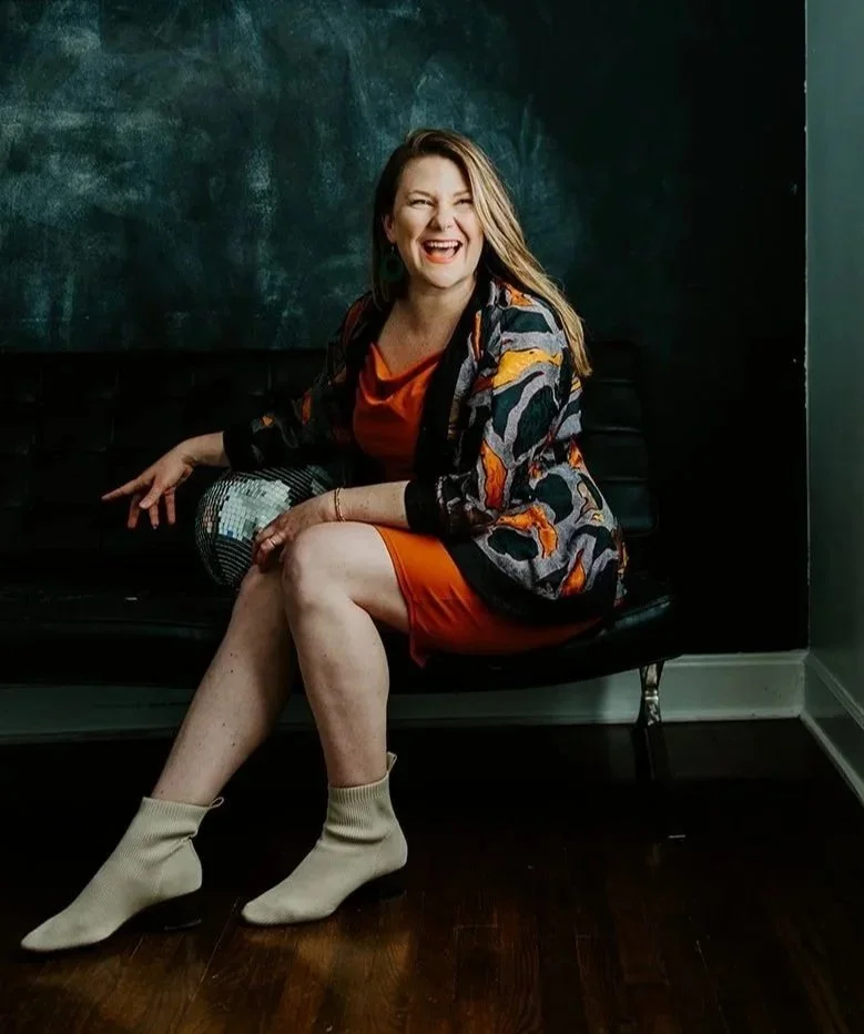 Woman sitting on a black bench, laughing, with a chalkboard behind her, wearing a colorful jacket over an orange dress and beige socks.