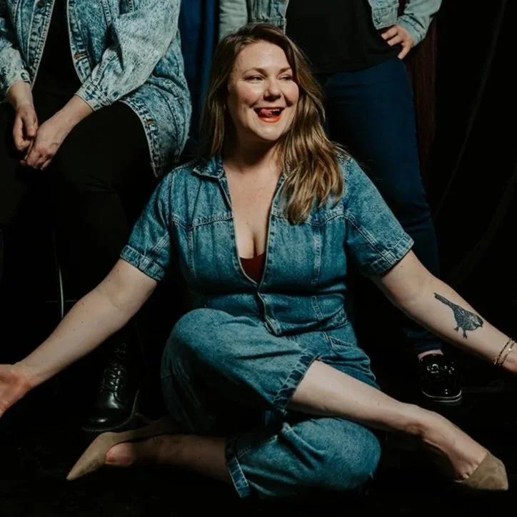 A woman in denim jumpsuit sitting on the floor with her legs crossed and arms extended. She has long hair, freckles, and is smiling with her tongue slightly out. She has a tattoo on her left forearm and is surrounded by others in denim and casual clothing.