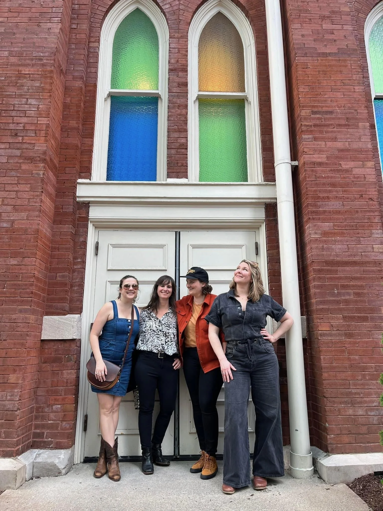 Four women standing in front of a white door with colorful stained glass windows on a brick building. They are smiling and looking cheerful.