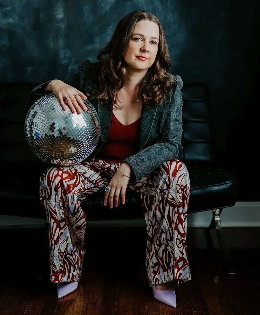 A woman with wavy brown hair sitting on a black leather couch holding a large disco ball in her left hand, dressed in a gray blazer, red top, and patterned pants, against a dark textured background.