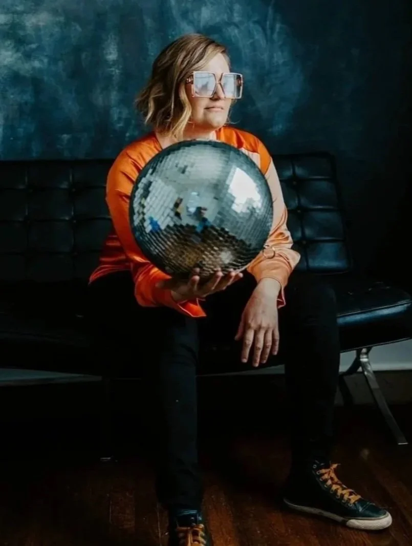 A woman with short blonde hair wearing large reflective sunglasses, an orange satin jacket, black pants, and patterned sneakers, sitting on a black bench against a blue textured wall, holding a large mirror ball.