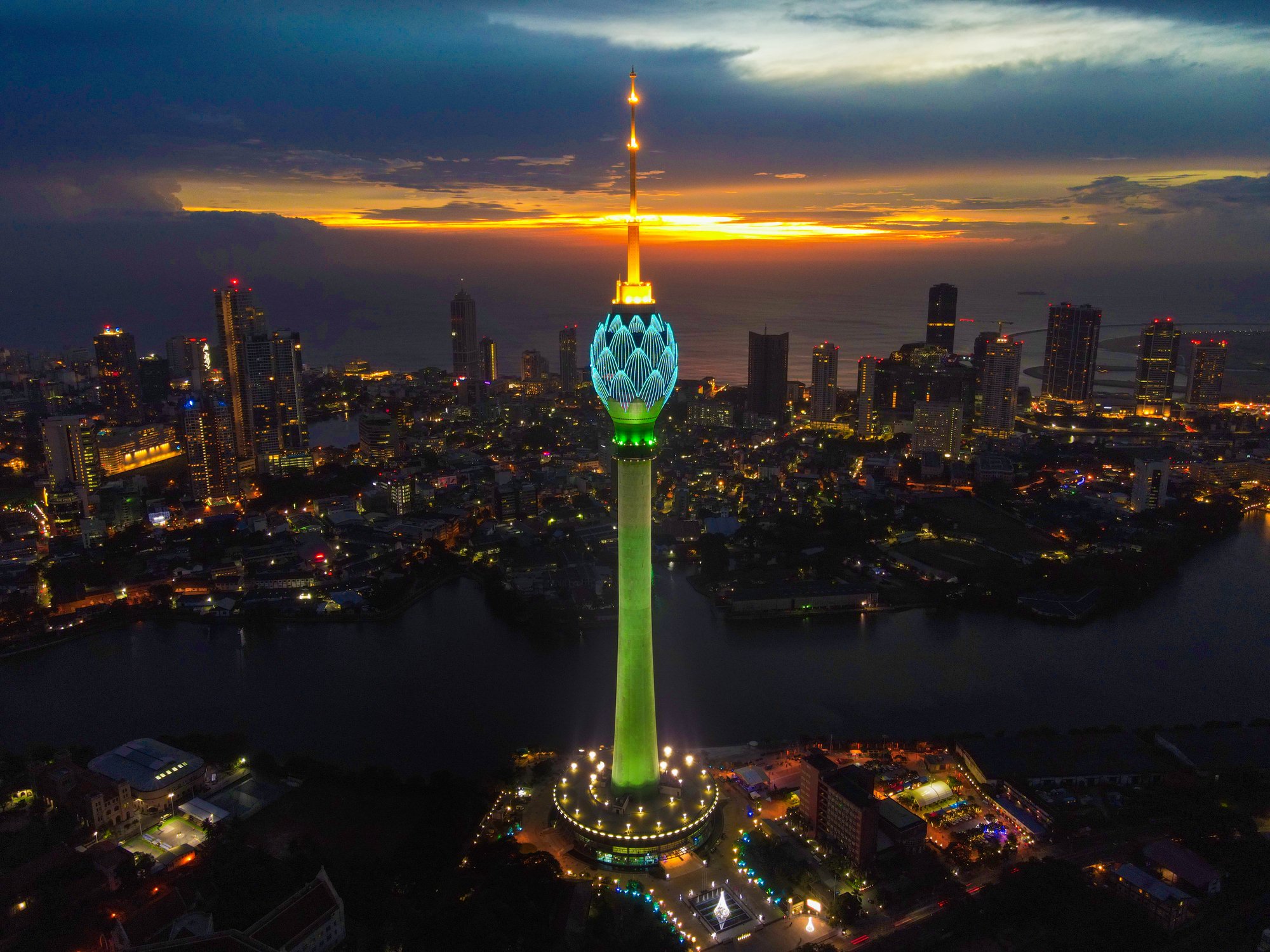 Aerial view of a tall illuminated tower at sunset in a cityscape, with colorful lights and surrounding buildings, water bodies, and an orange sky.