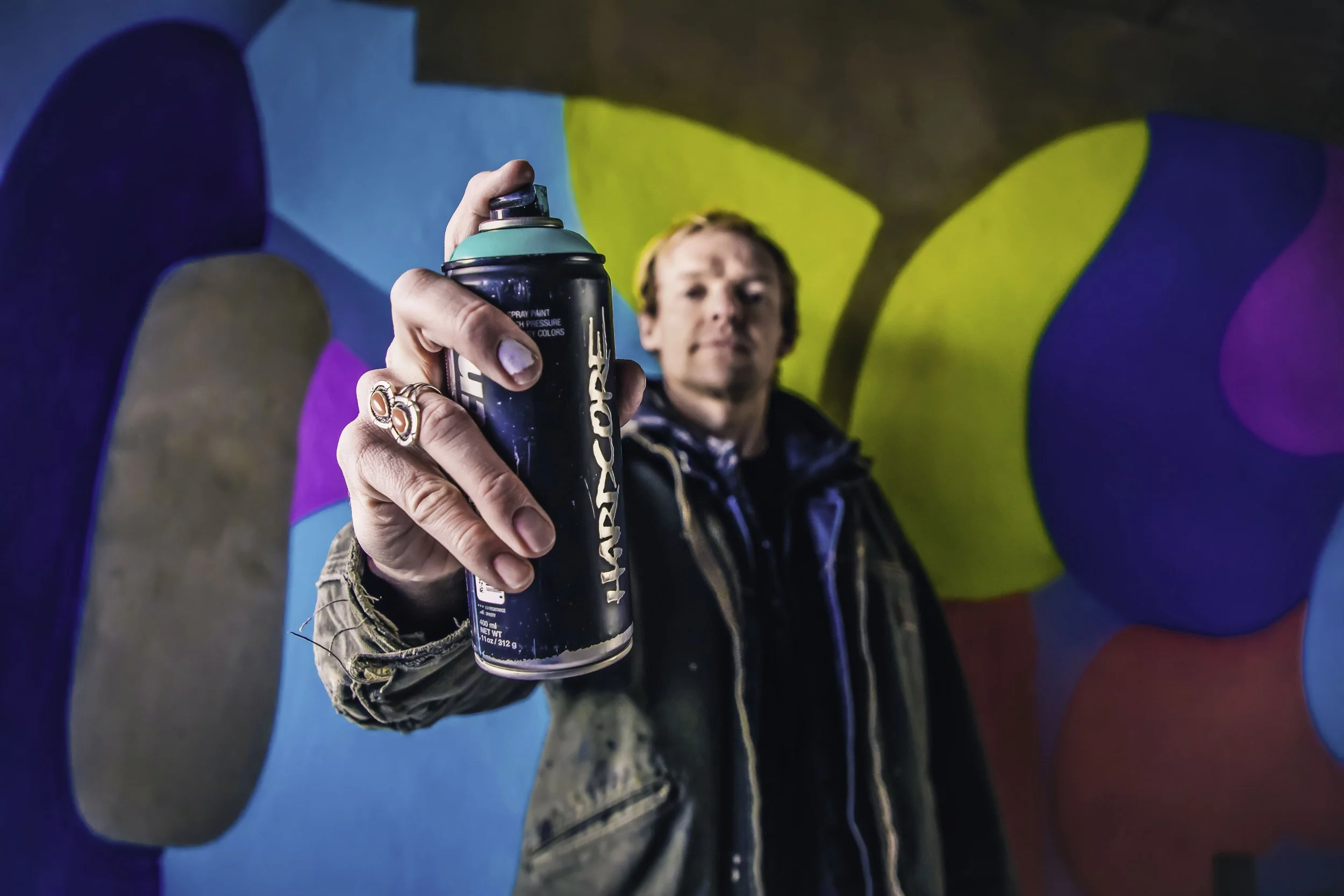 A person holding a can of spray paint towards the camera, with a colorful abstract mural in the background.
