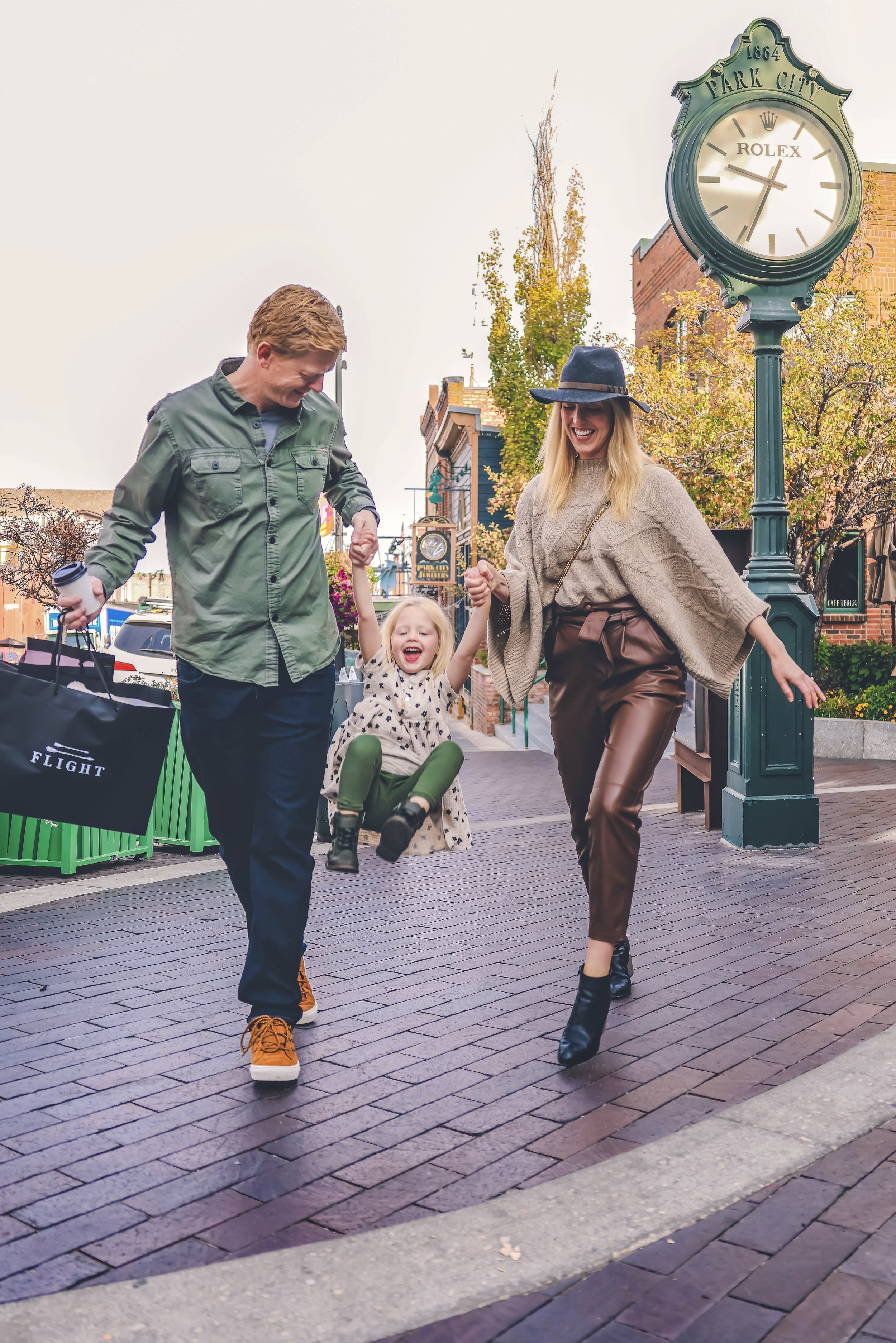 A man, a woman, and a young girl are walking together on a brick sidewalk in a downtown area. The man is holding shopping bags, the girl is riding a small scooter, and they are all smiling and holding hands, enjoying their time outside. There is a st