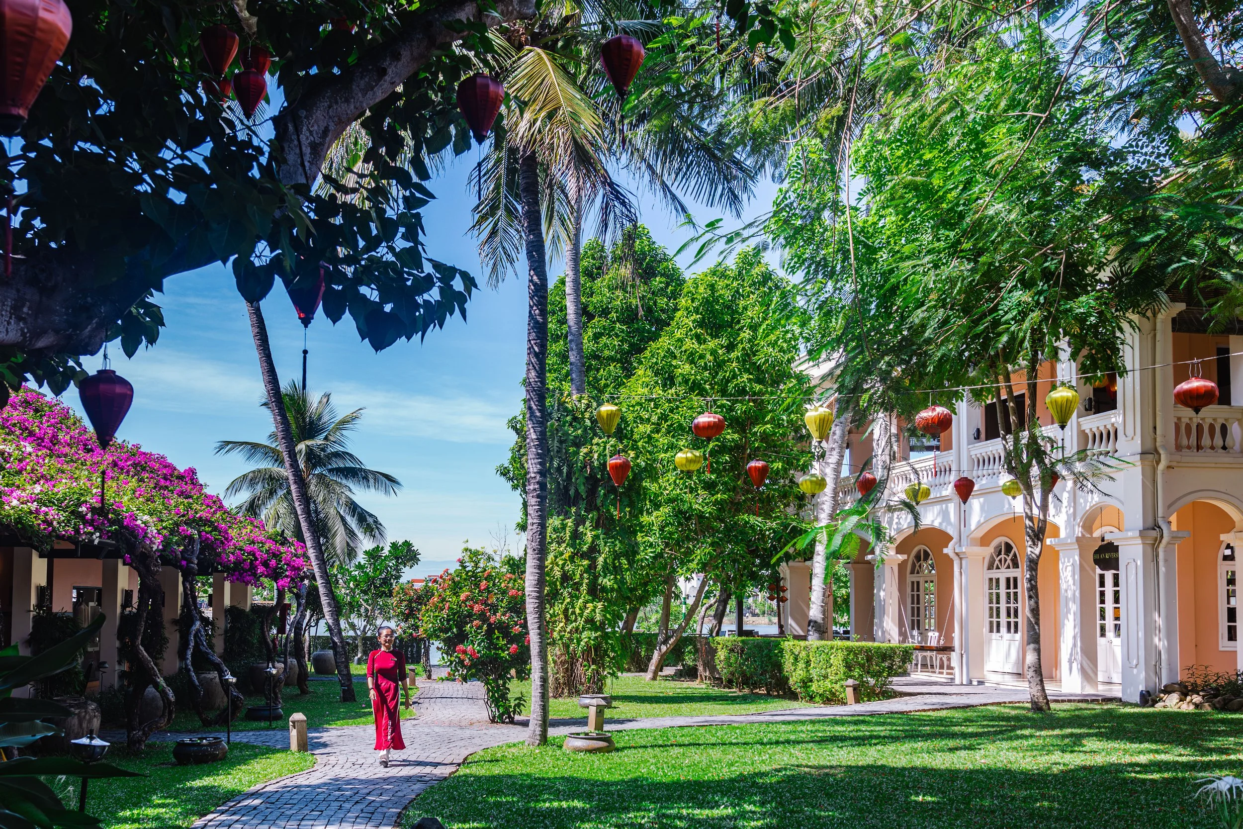 Colorful lanterns hanging over a lush green courtyard with trees, flowers, and a woman in a red dress walking on a cobblestone path near a cream-colored building with arches.