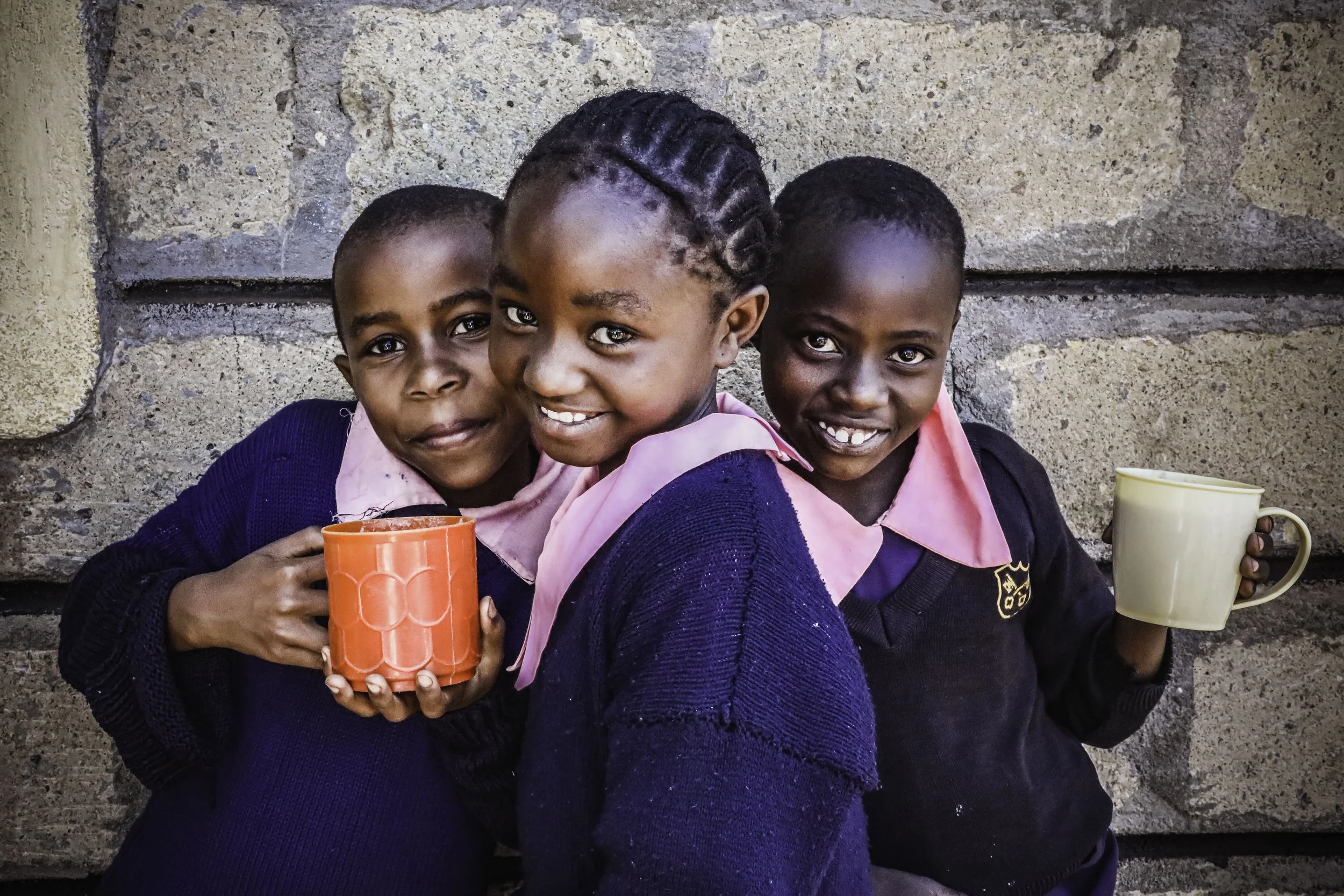 Three young African children smiling while holding mugs, sitting against a stone wall.
