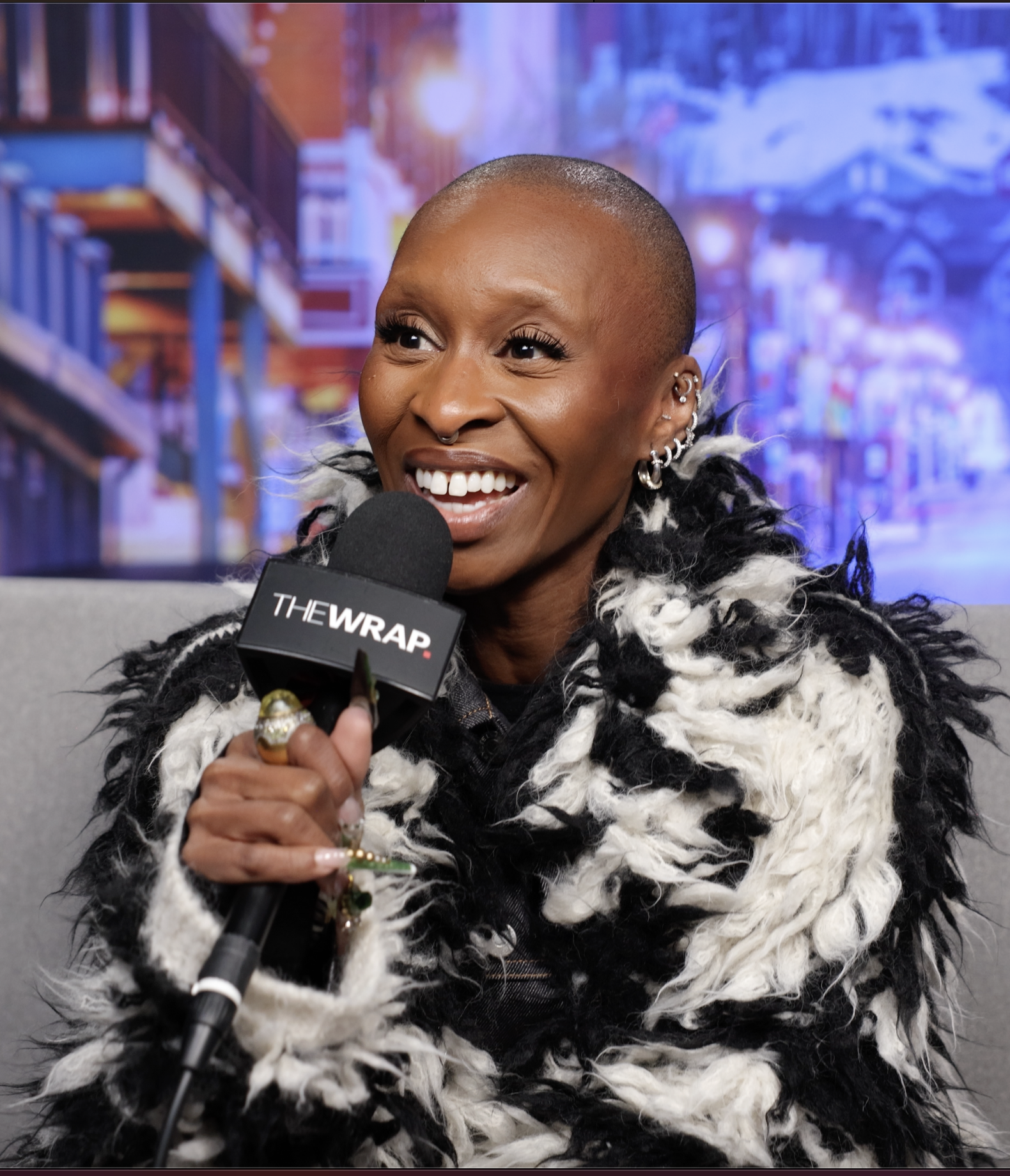 Close-up of a laughing woman holding a microphone with 'THE WRAP' written on it, with a blurred cityscape background.