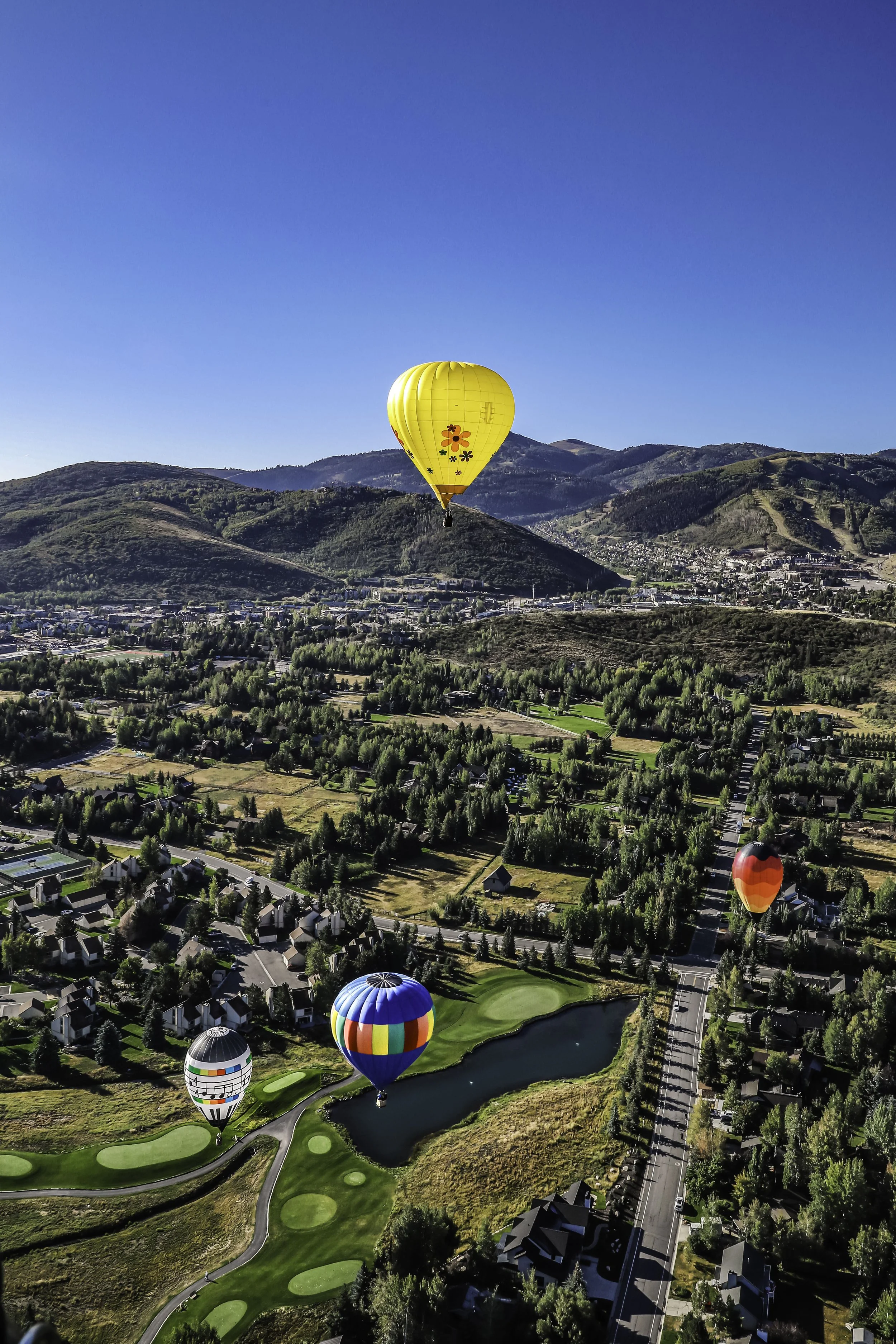 Multiple colorful hot air balloons flying over a scenic landscape of hills, a golf course, residential houses, and a lake on a clear blue day.