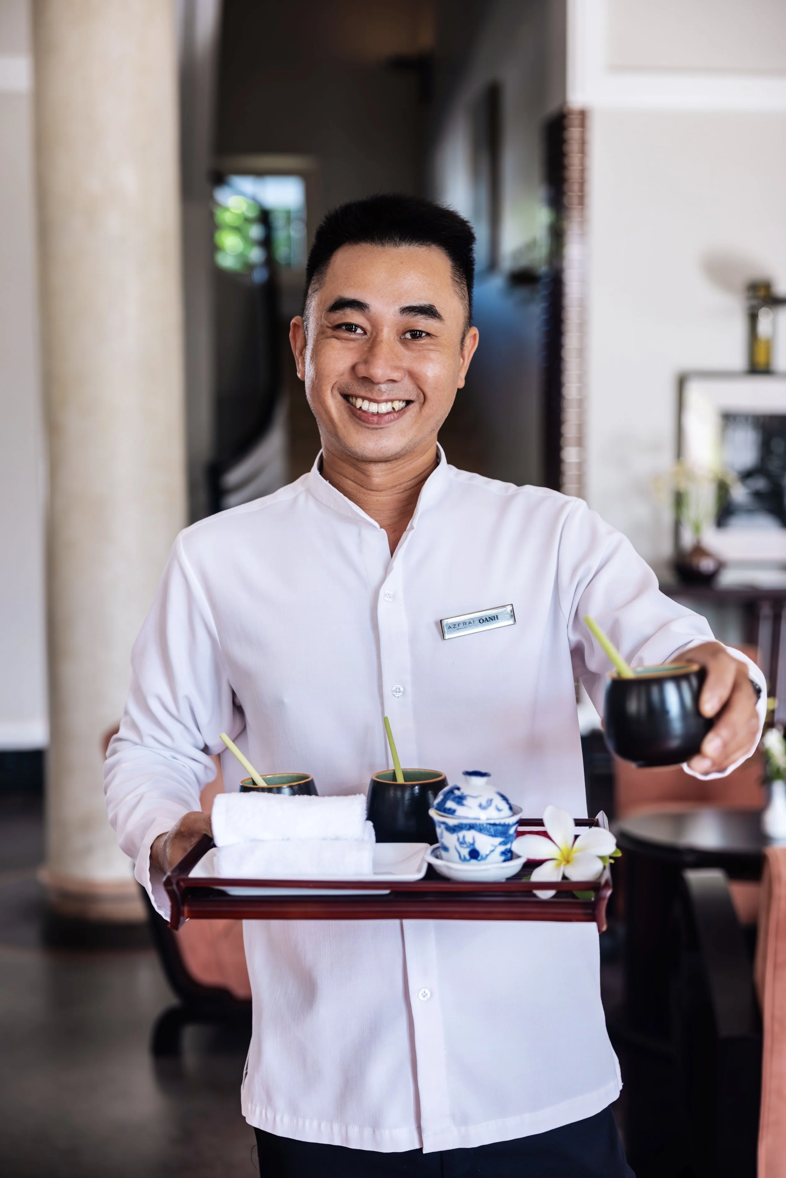 A smiling waiter in a white uniform holding a tray with drinks and a towel inside a restaurant.