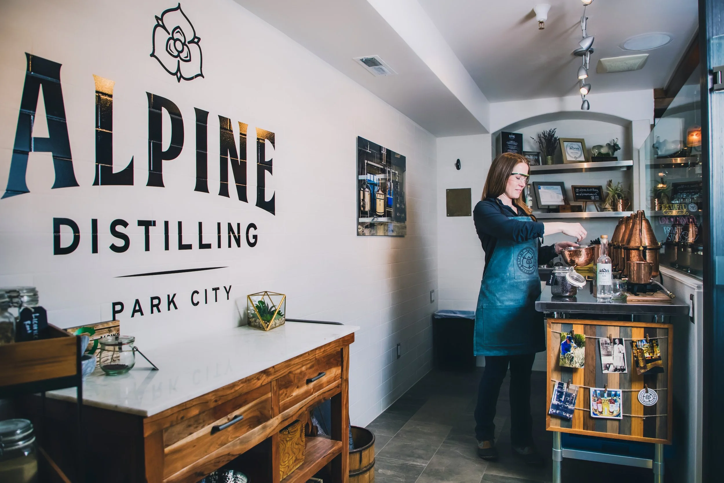 Interior of a distillery shop with a woman behind the counter, shelves with bottles and souvenirs, and a wall with a large logo reading 'ALPINE DISTILLING'.