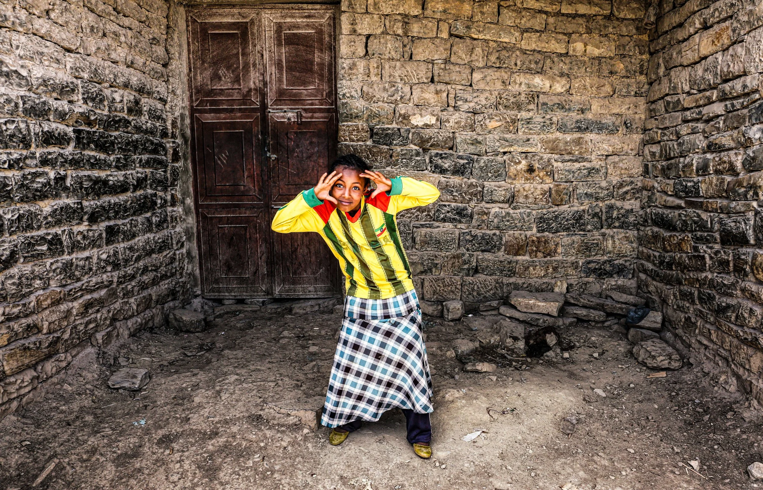 A young boy standing in front of a weathered stone wall with a dark wooden door, wearing a colorful striped long-sleeve shirt and a plaid skirt, holding his hands to his head in a playful gesture.
