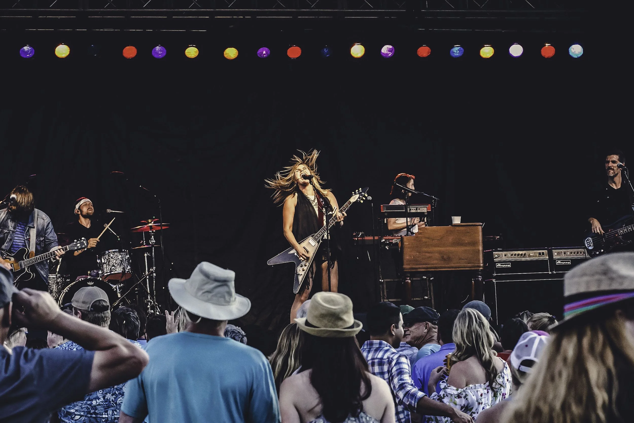 A live band performing on stage with members playing guitars, drums, keyboard, and singing into microphones, audience watching in front, some wearing hats.