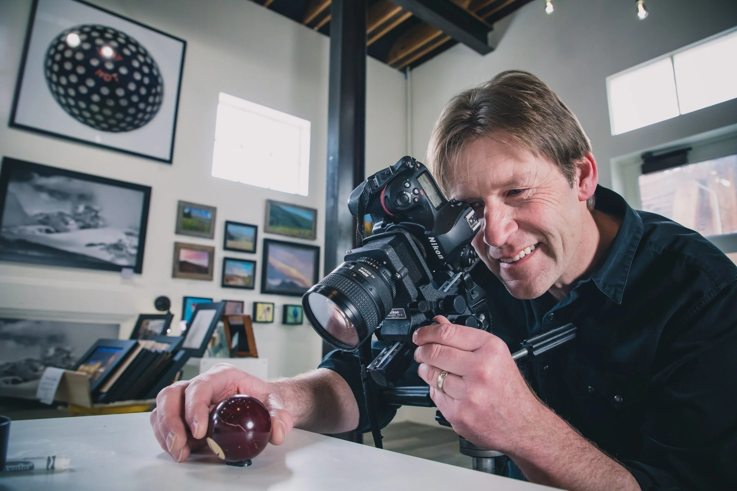 A man with a camera taking a photograph of a maroon apple on a white table in an art gallery or studio, with framed artwork on the walls behind him.