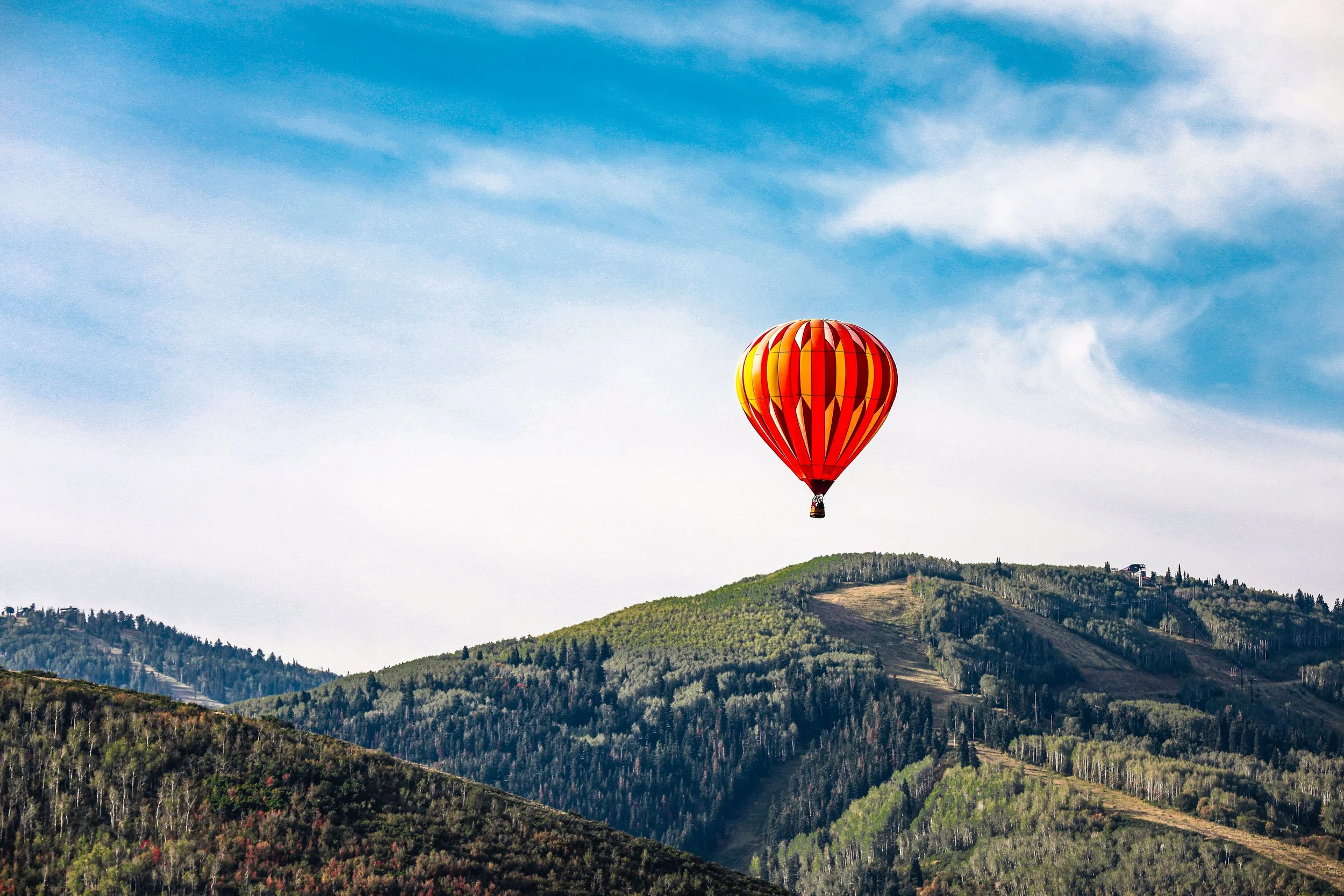 A hot air balloon with red, orange, yellow, and white patterns floating in a partly cloudy sky above green rolling hills and trees.