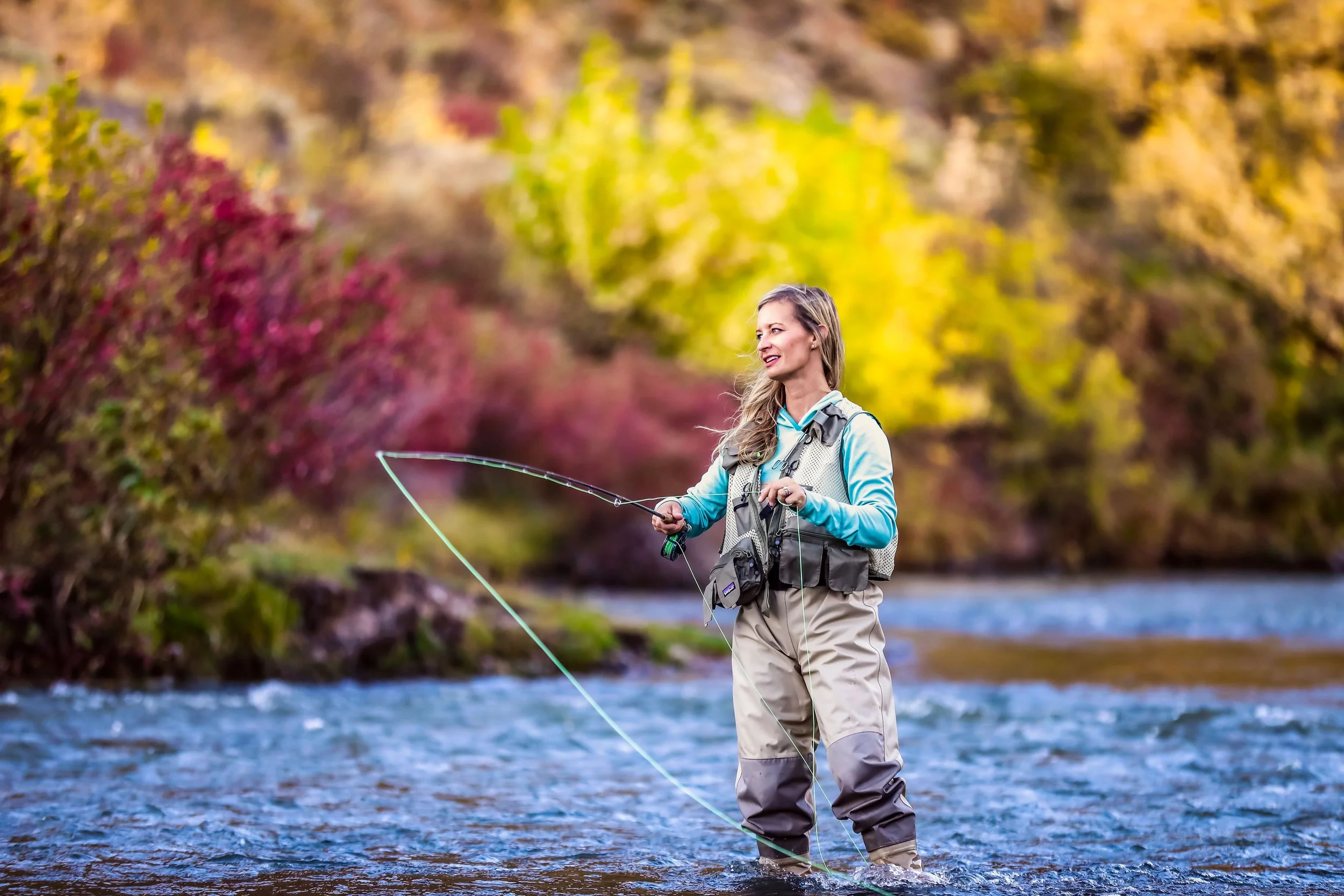 A woman fishing in a river surrounded by trees with colorful autumn leaves.