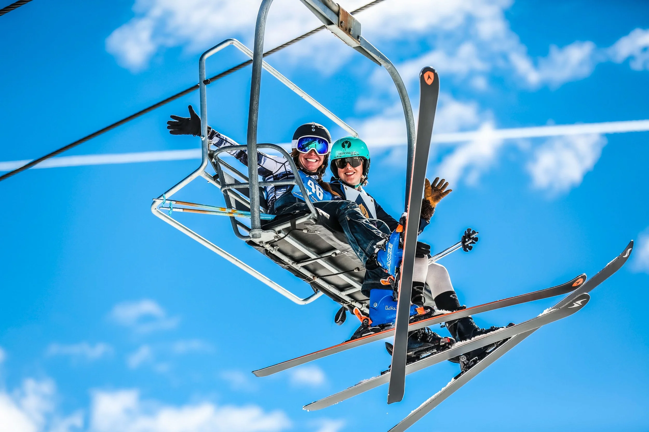 Two women smiling and waving from a ski lift, wearing ski helmets, goggles, and winter gear, with blue sky and clouds in the background.