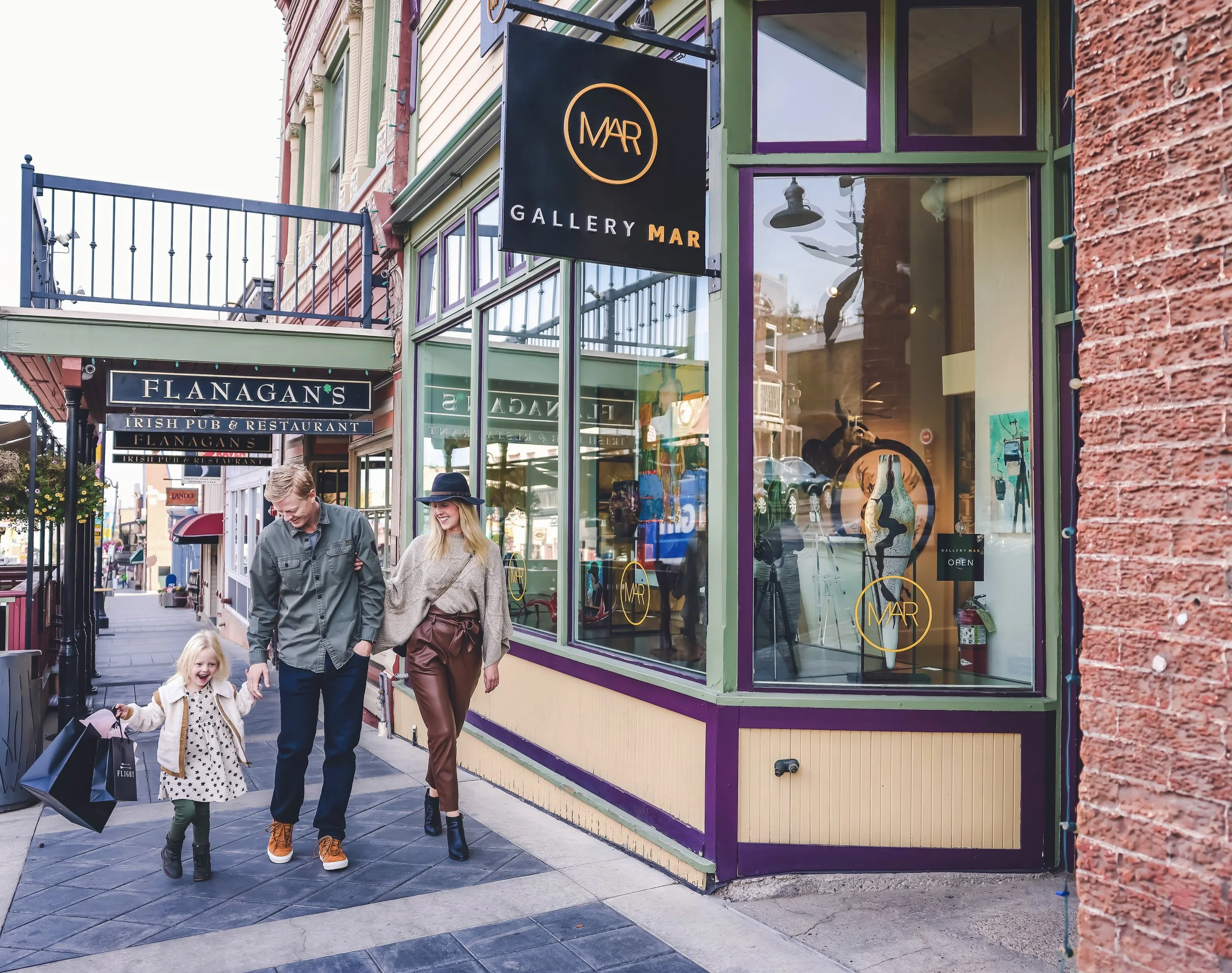 A man, woman, and young girl walking on a city sidewalk in front of a storefront with large glass windows. The storefront has a sign reading 'GALLERY MAR' and is decorated with colorful trim. The group is smiling, and the girl is holding shopping bag