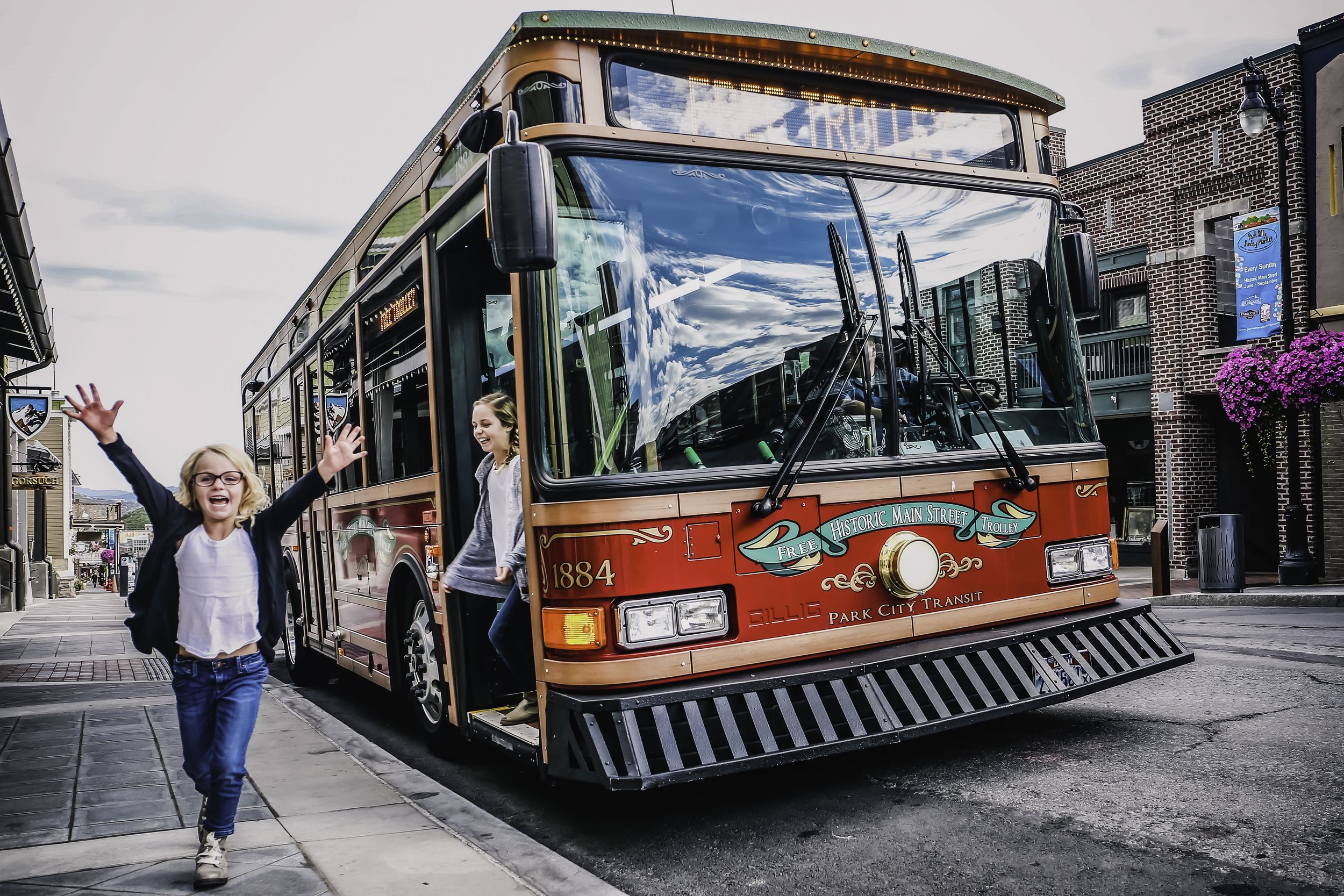 Two children waving as they walk away from a red trolley streetcar on a city street. The trolley has a sign displaying 'FRISCO' and the words 'Park City Transit' on its front. The street is lined with shops and buildings, some with flower baskets.