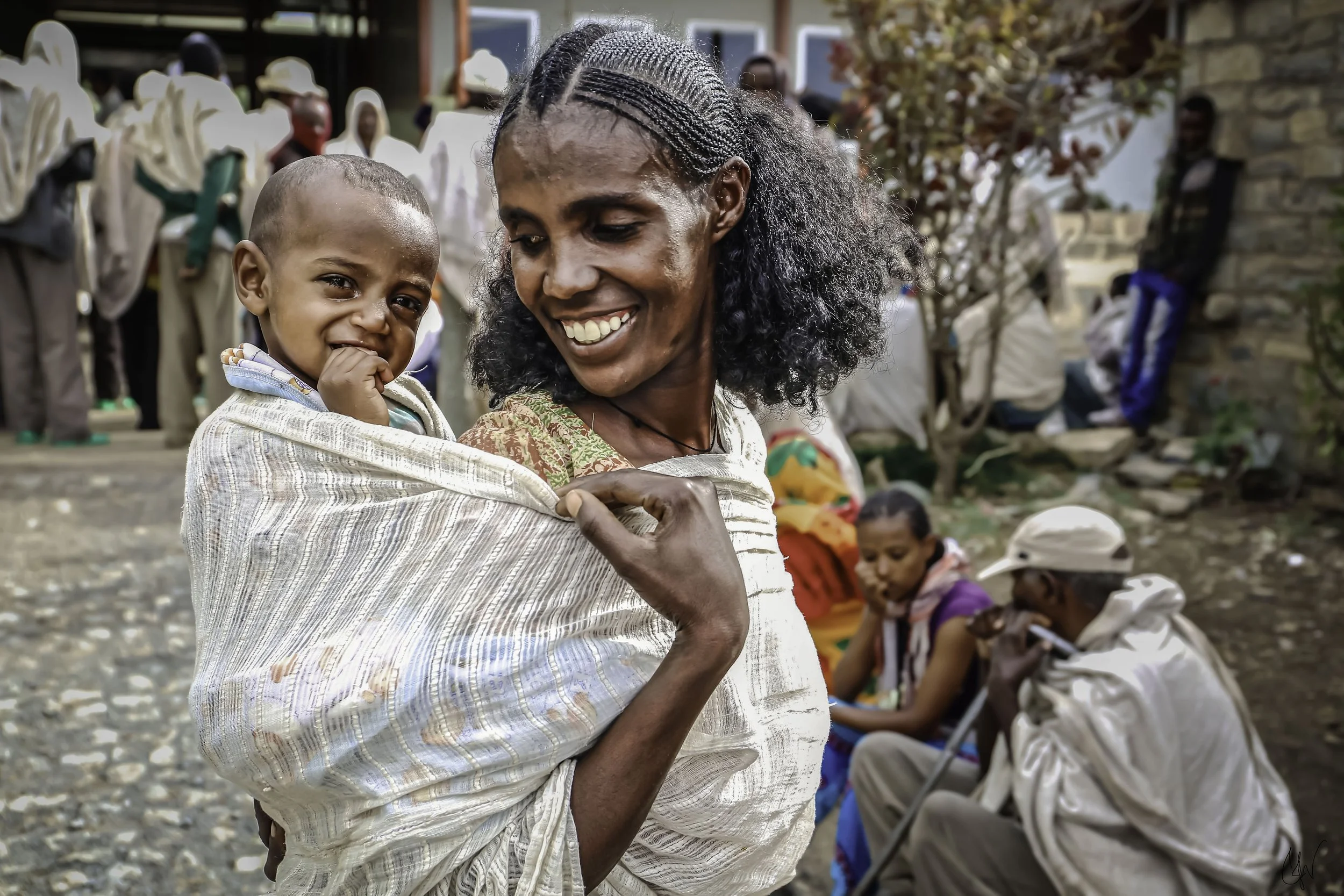 A smiling woman with braided hair holding a young boy who appears to be upset, among other people sitting and standing outdoors in a community setting.