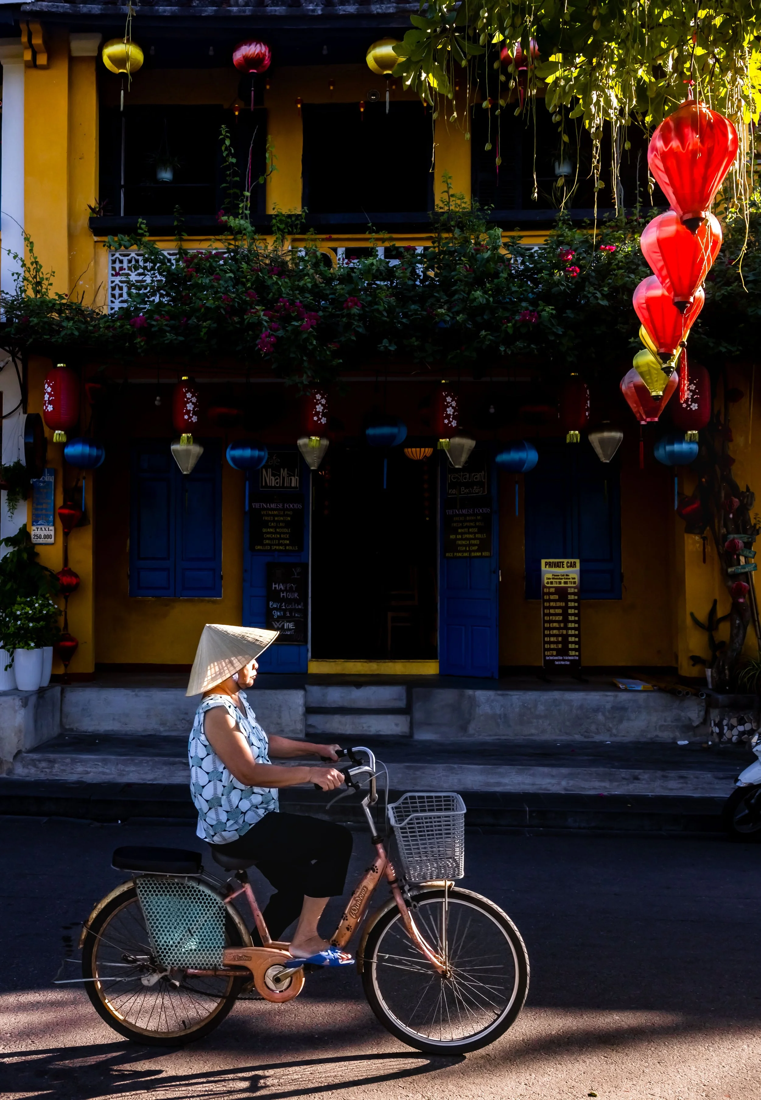 A woman wearing a conical Asian hat and a sleeveless floral shirt is riding a pink bicycle with a basket on the front. She is moving along a street in front of a yellow building with blue doors and colorful hanging lanterns. There is greenery on the 