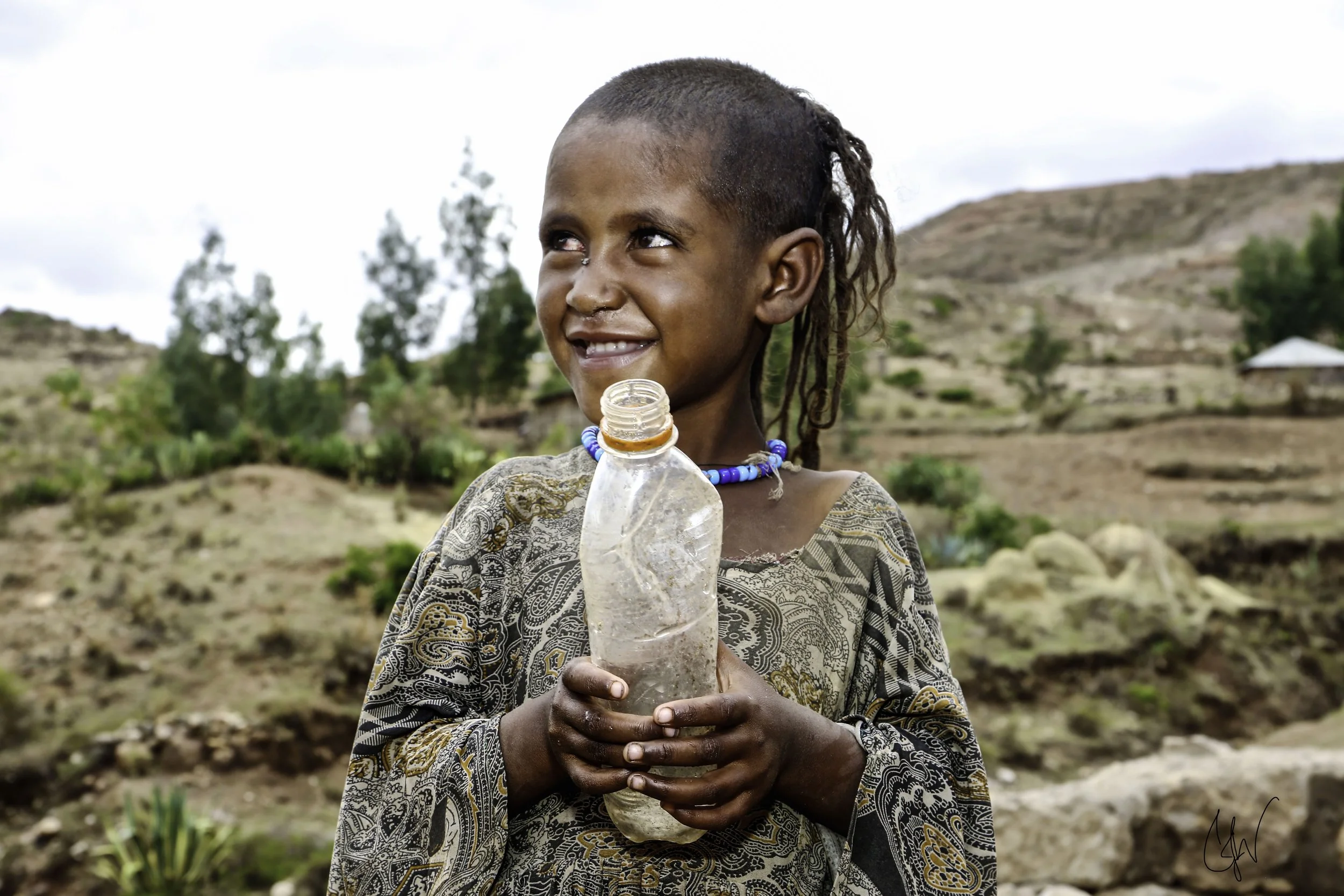 A young girl with braided hair, wearing a patterned dress and a blue beaded necklace, holding a clear plastic water bottle outdoors in a rural setting with hills and sparse trees in the background.