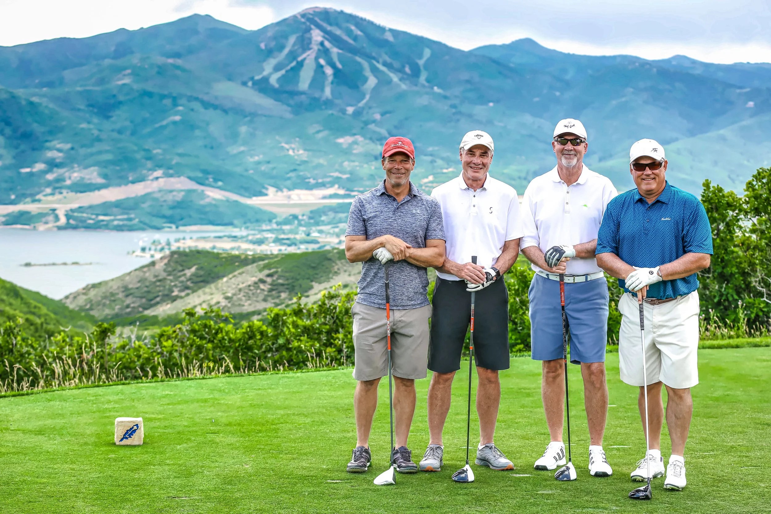 Four men standing on a golf course with mountains in the background, holding golf clubs and smiling.