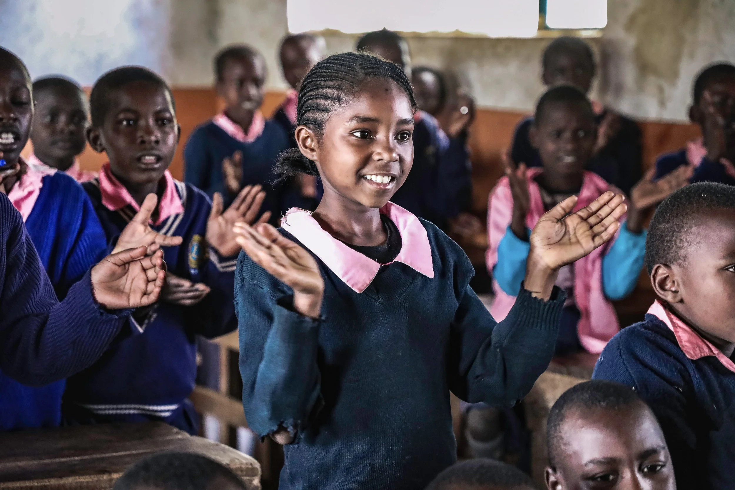 Young children in school uniforms participating in a classroom activity, standing with hands raised, inside a rustic classroom with a window.