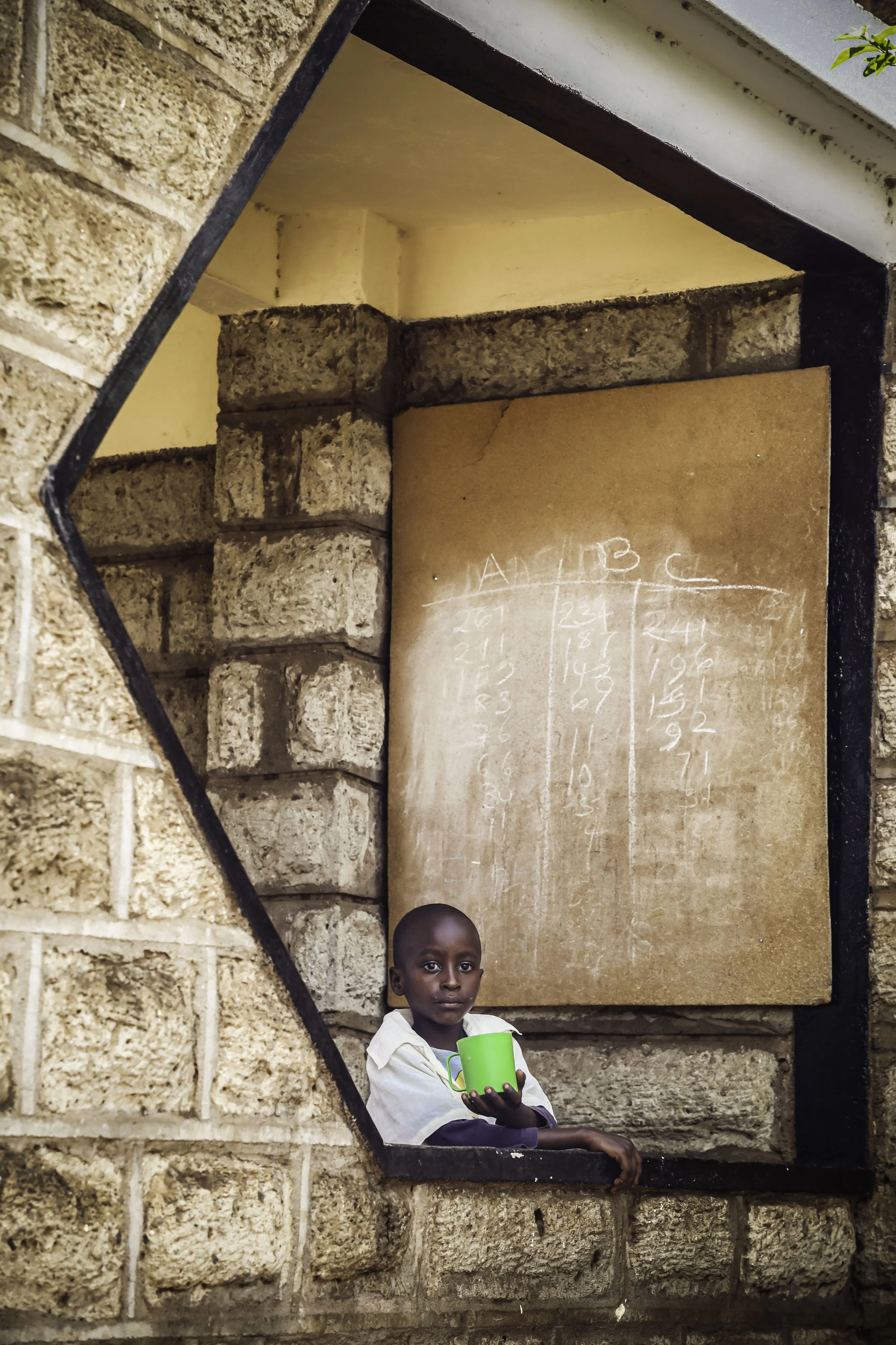 A young boy holding a green cup, standing in front of a brick wall with a chalkboard behind him, in an outdoor classroom setting.