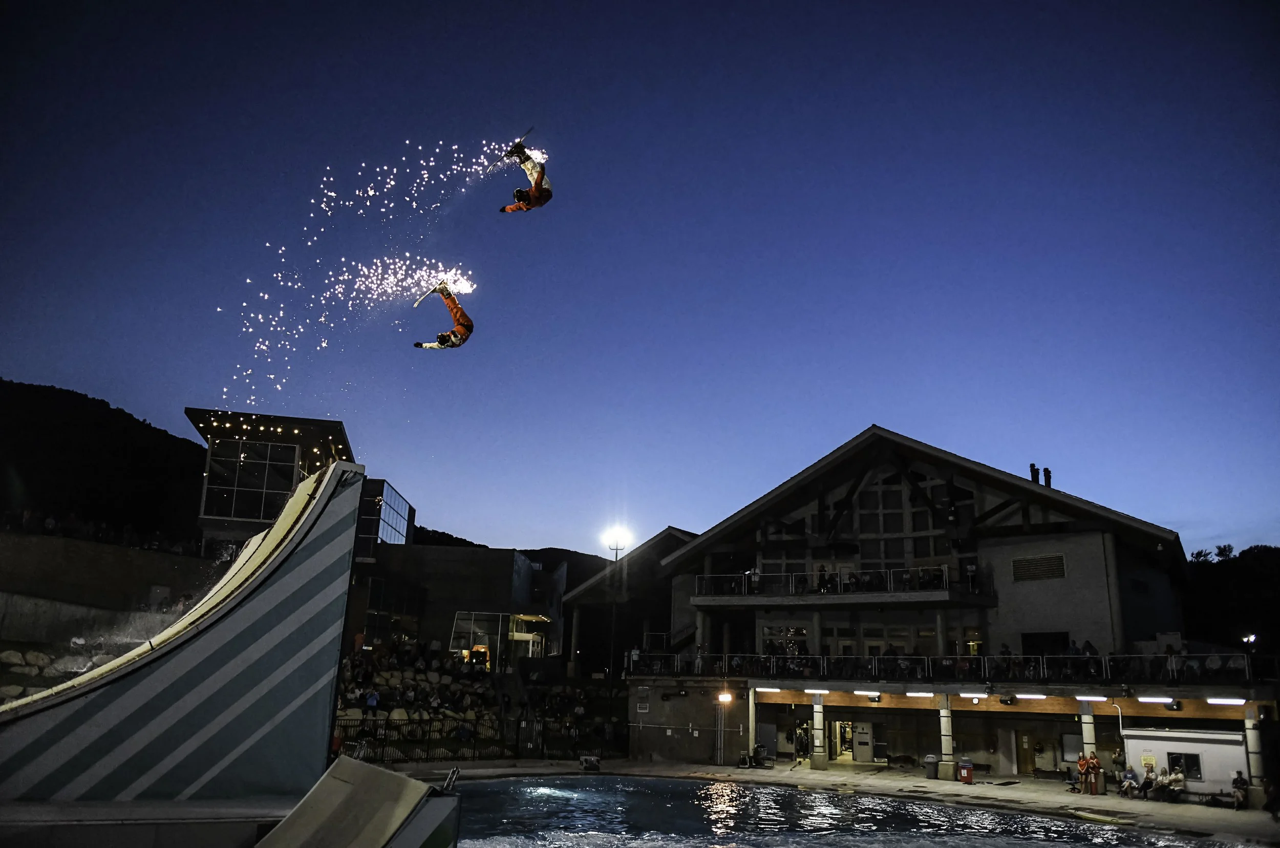 Two snowboarders performing aerial tricks on a halfpipe at a ski resort during dusk, with a lit building and audience below.